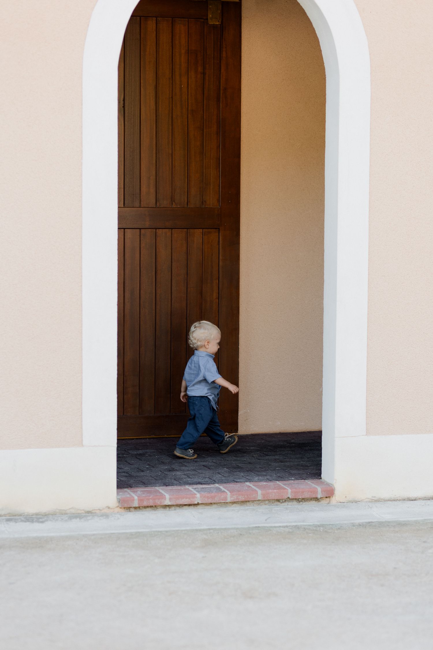 A small child peeks around the corner of a beige building with arched doorways and dark wooden doors.