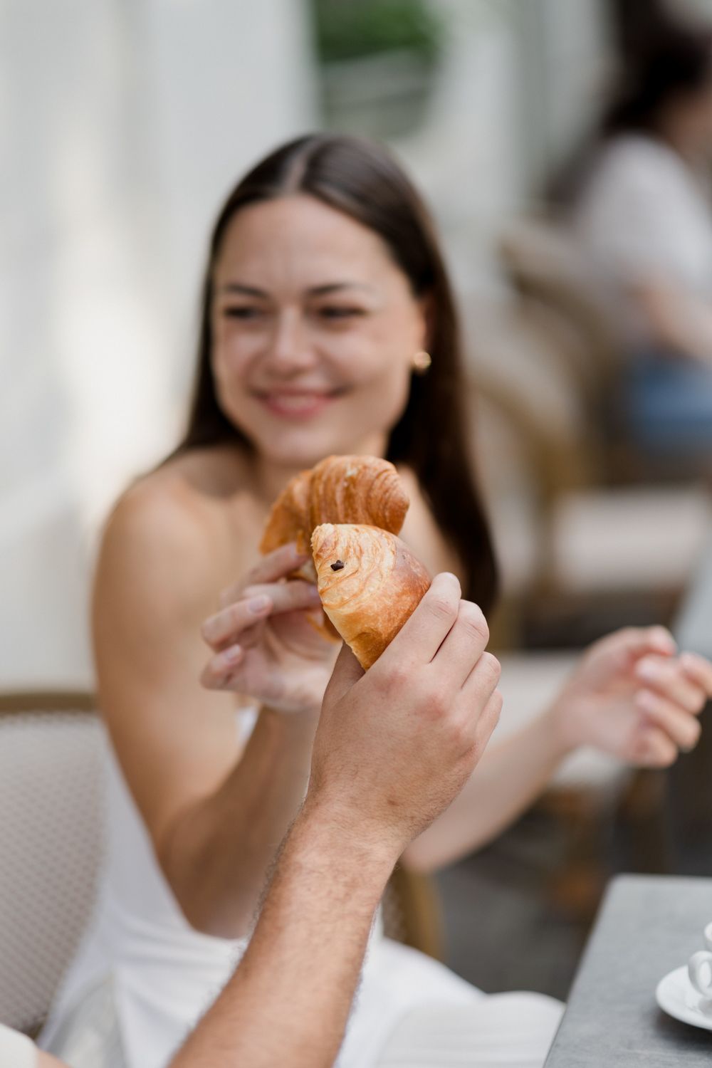 Someone's hand holds up a freshly baked croissant in a restaurant setting while another person smiles in the background.