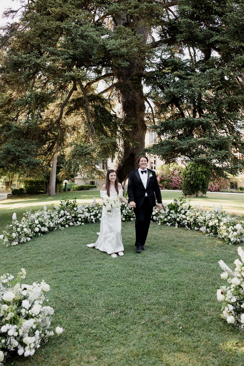 A couple in wedding attire stands beside a curved path lined with elegant white floral arrangements in a garden setting.