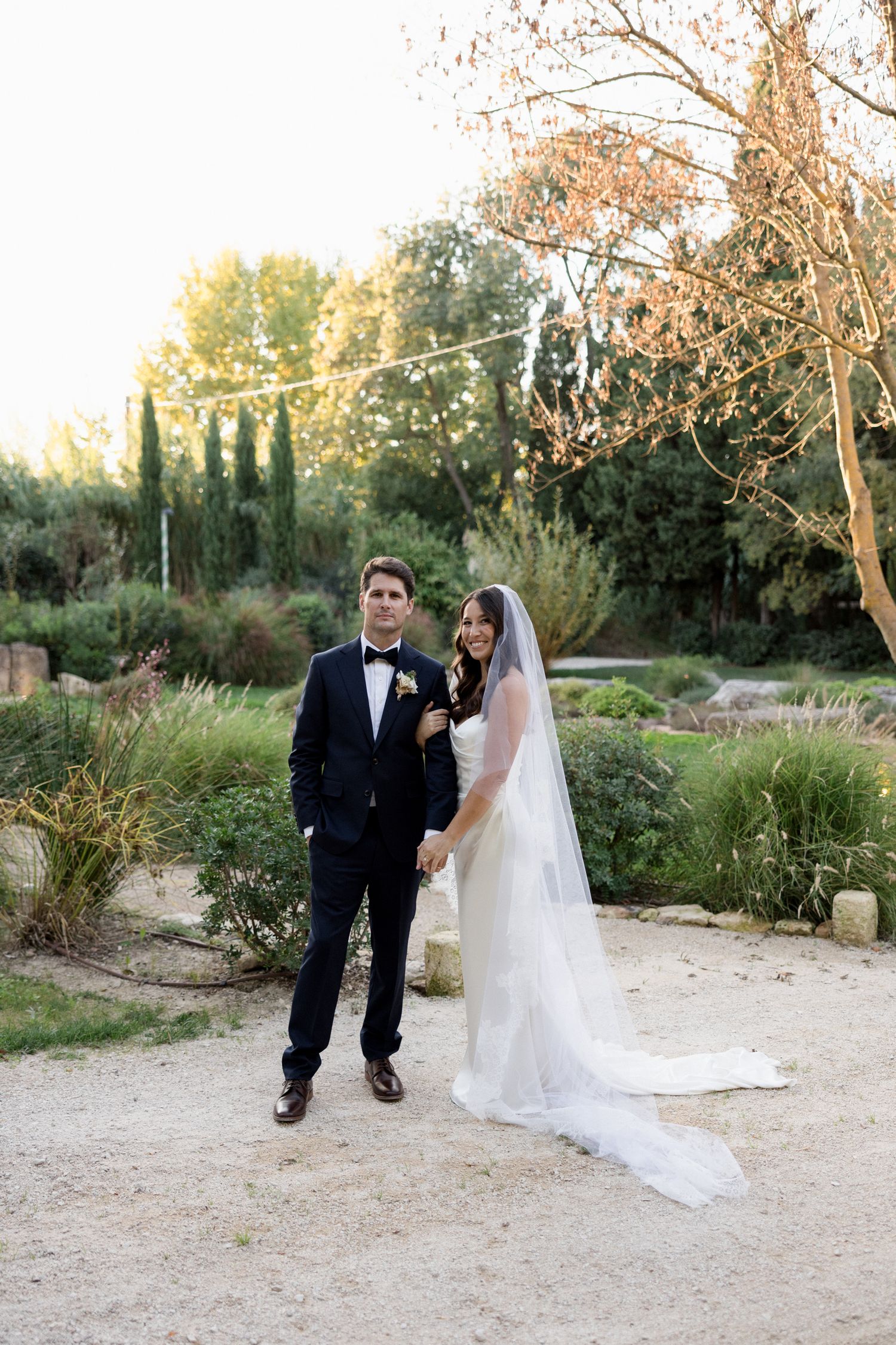 A couple in formal wedding attire poses together in a garden setting with natural lighting at sunset.