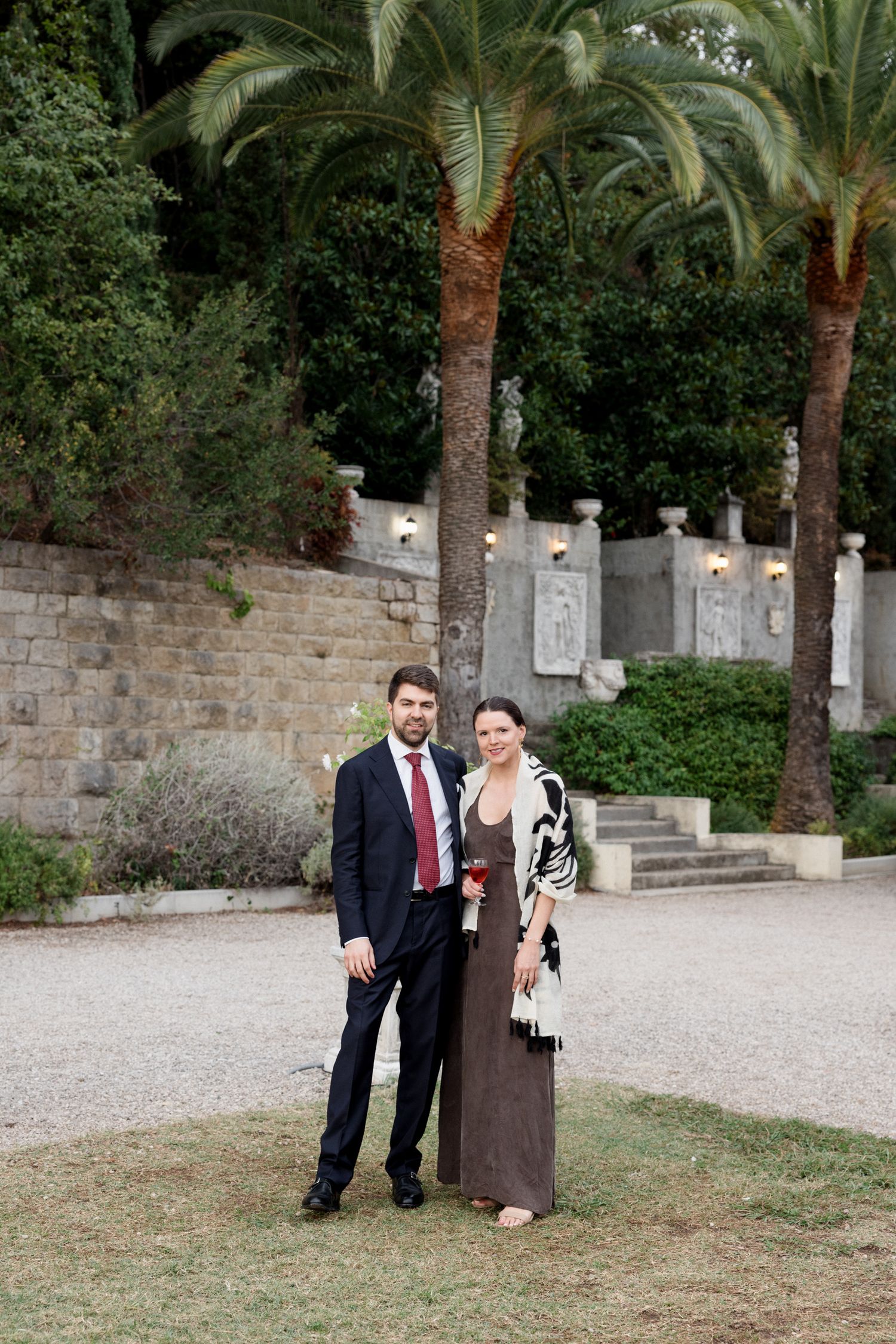 A couple poses together in a garden setting with palm trees and stone walls in the background.