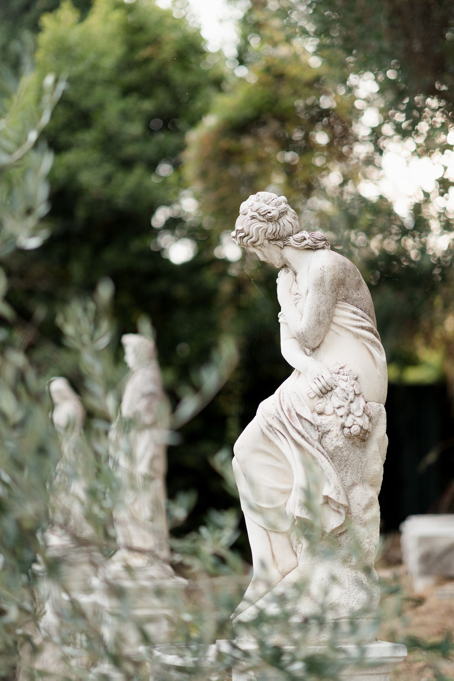 A series of white marble cherub statues displayed in a garden setting with soft bokeh effects and natural lighting.