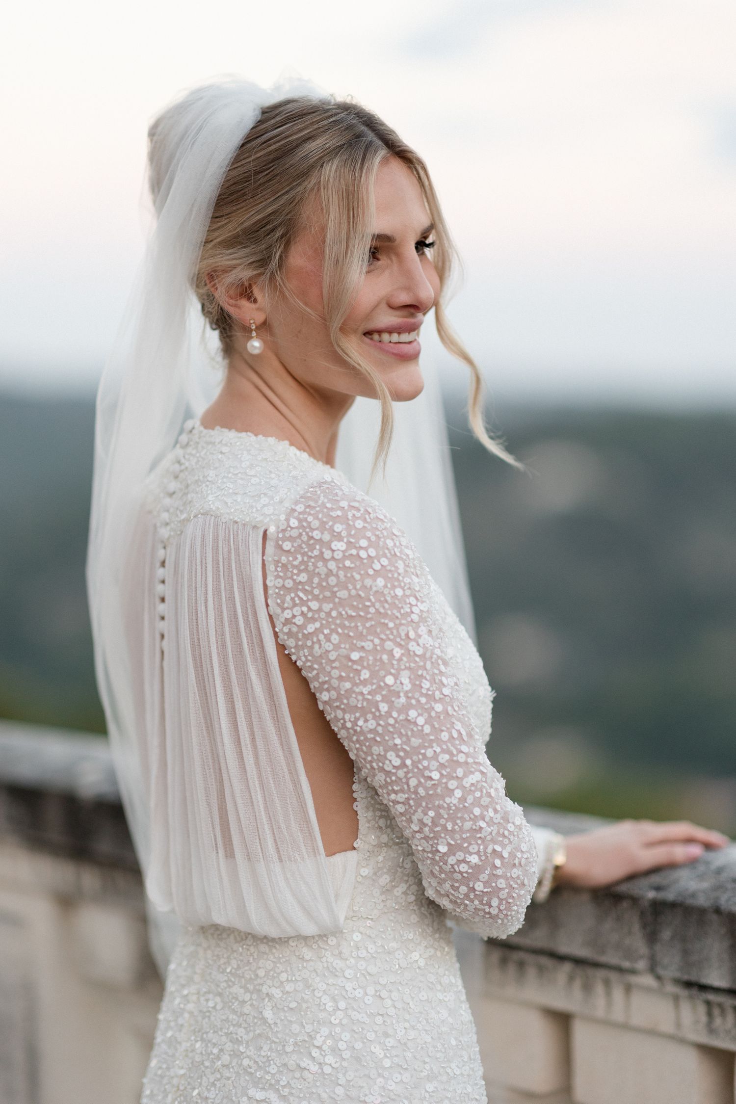 Elegant bride in long-sleeved white beaded wedding dress with veil poses against stone balcony in outdoor setting.