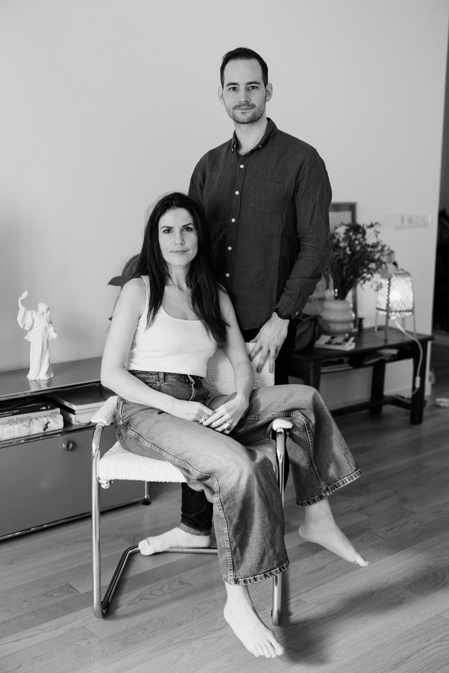 Black and white photo of a couple posing indoors, woman seated in chair, man standing behind her.