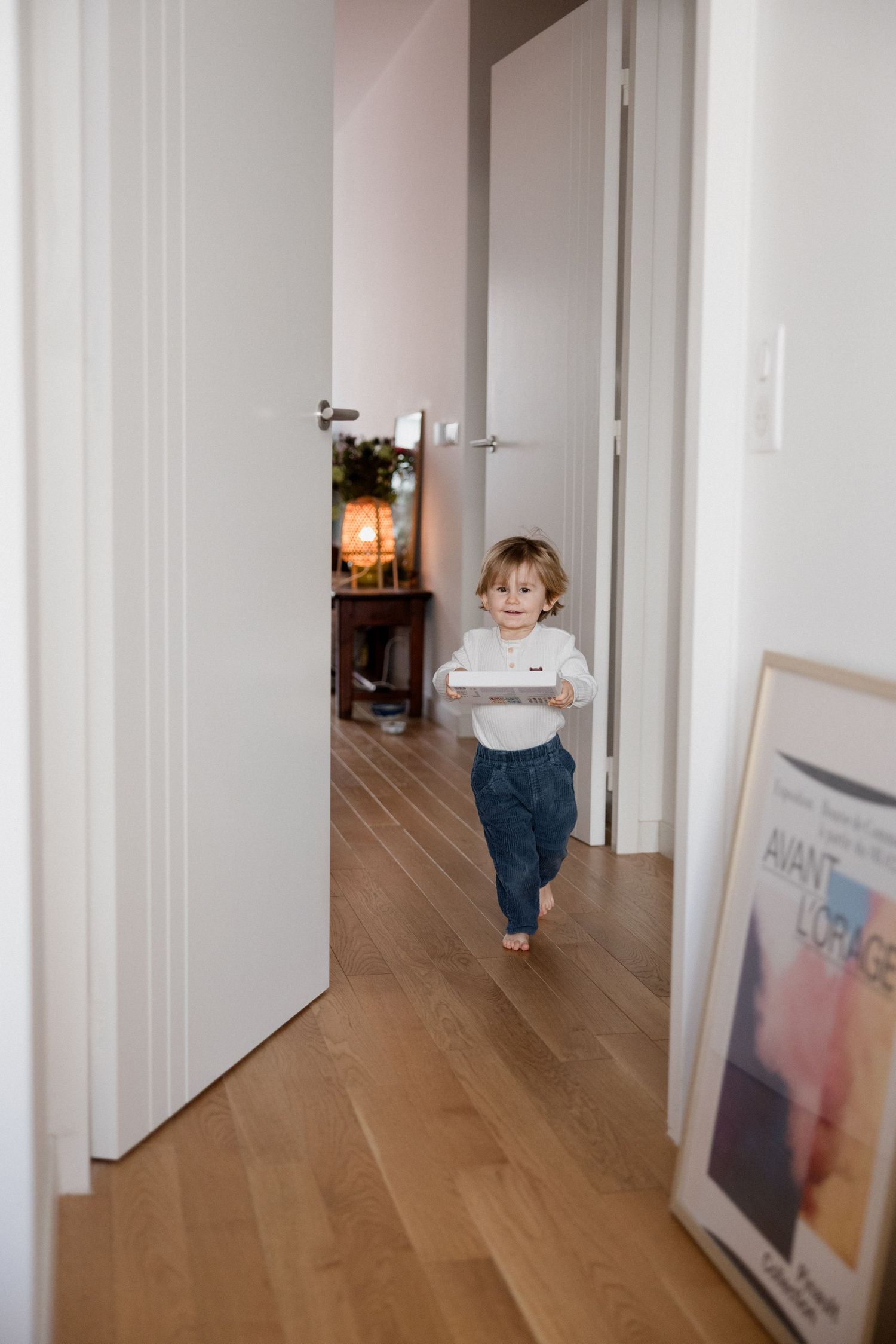 Toddler boy running barefoot through bright white hallway carrying a tablet in modern apartment.