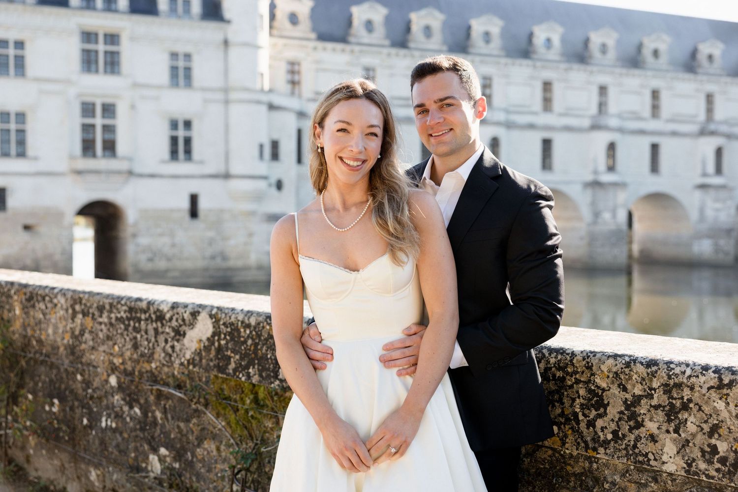 Smiling couple in wedding attire poses before Château de Chenonceau, bride in white dress with pearls, groom in black suit.