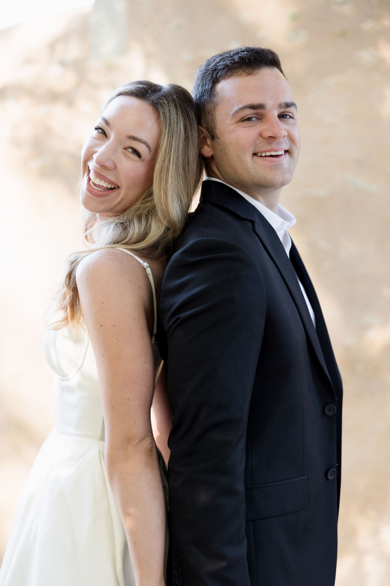 Smiling couple in wedding attire standing back-to-back, woman in white dress and man in black suit.