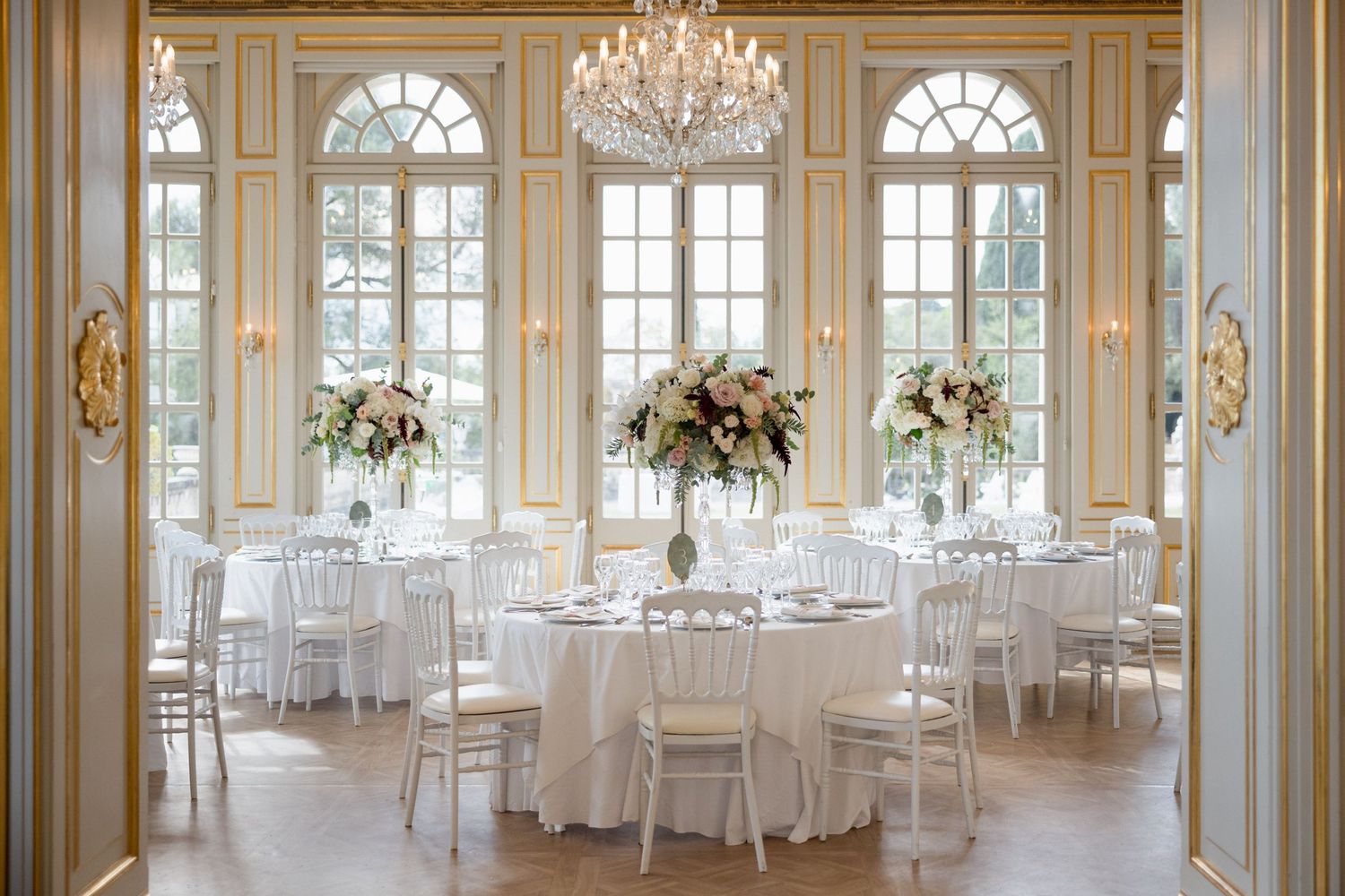 Elegant ballroom with crystal chandeliers, arched windows, ornate gold trim and white-clothed round tables for formal events. mariagegrasse