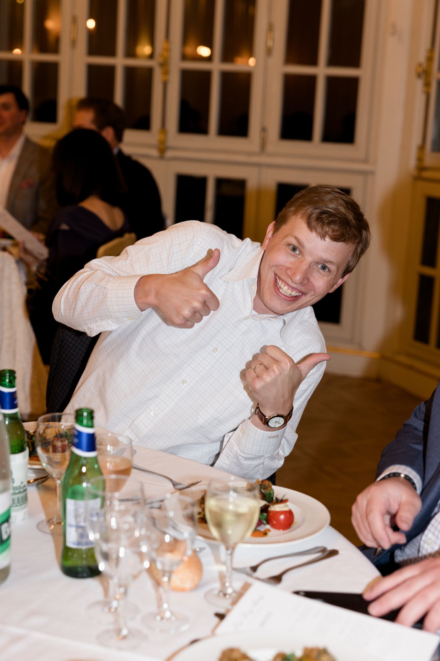 Dinner party guests in white shirts laughing and socializing around a table with wine bottles and glasses.