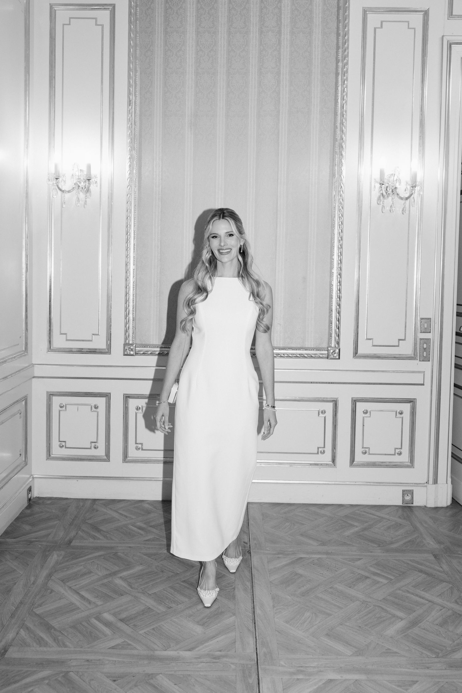 Elegant black and white photo of a person in a sleek white column dress standing in an ornate room with wall sconces. destination wedding fr