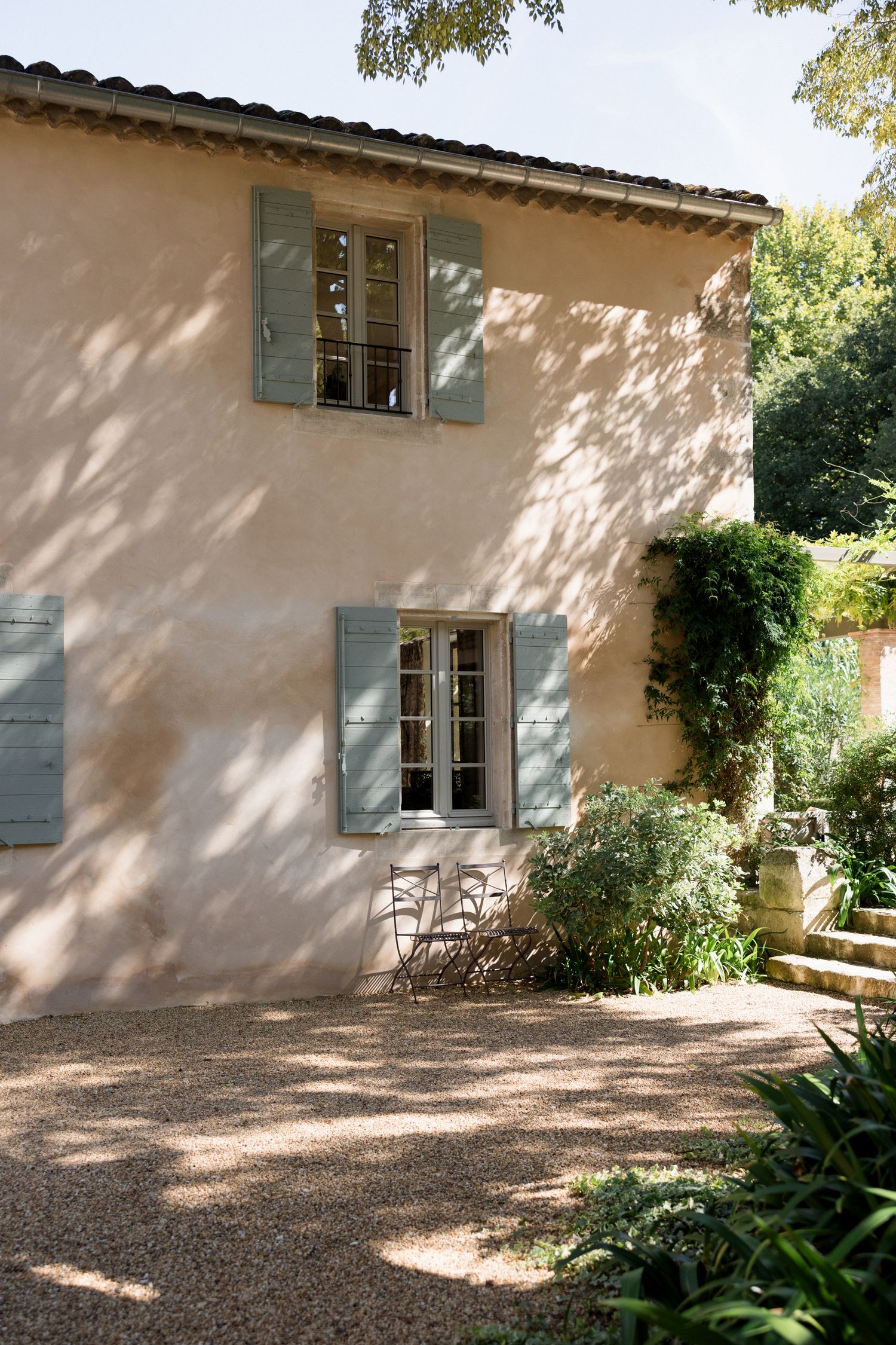 Mediterranean villa exterior with pale stucco walls, blue shuttered windows and gravel courtyard in dappled sunlight. Saint Remy de Provence
