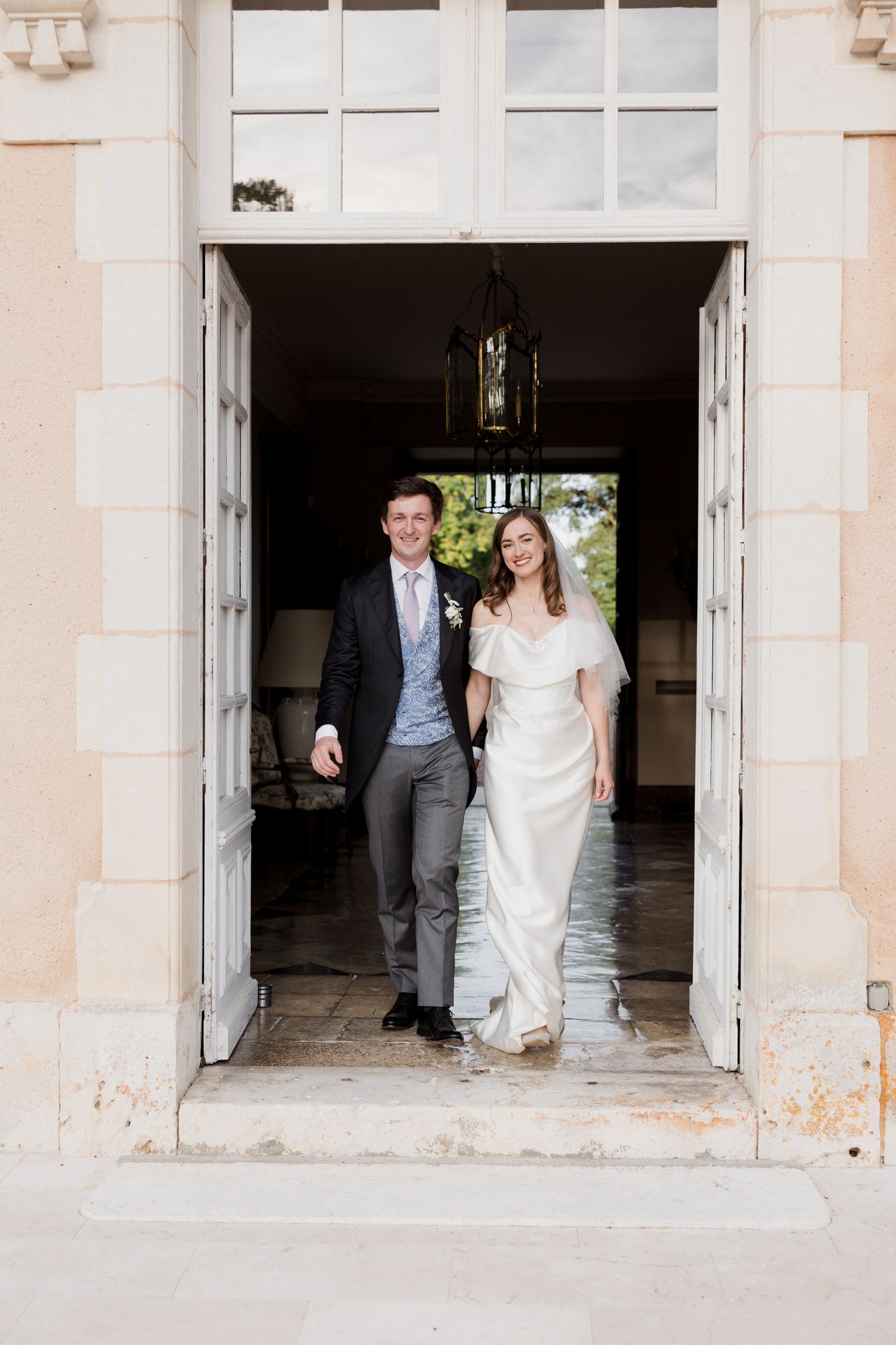 A couple in wedding attire walks through a grand stone doorway with white doors and a hanging lantern above.