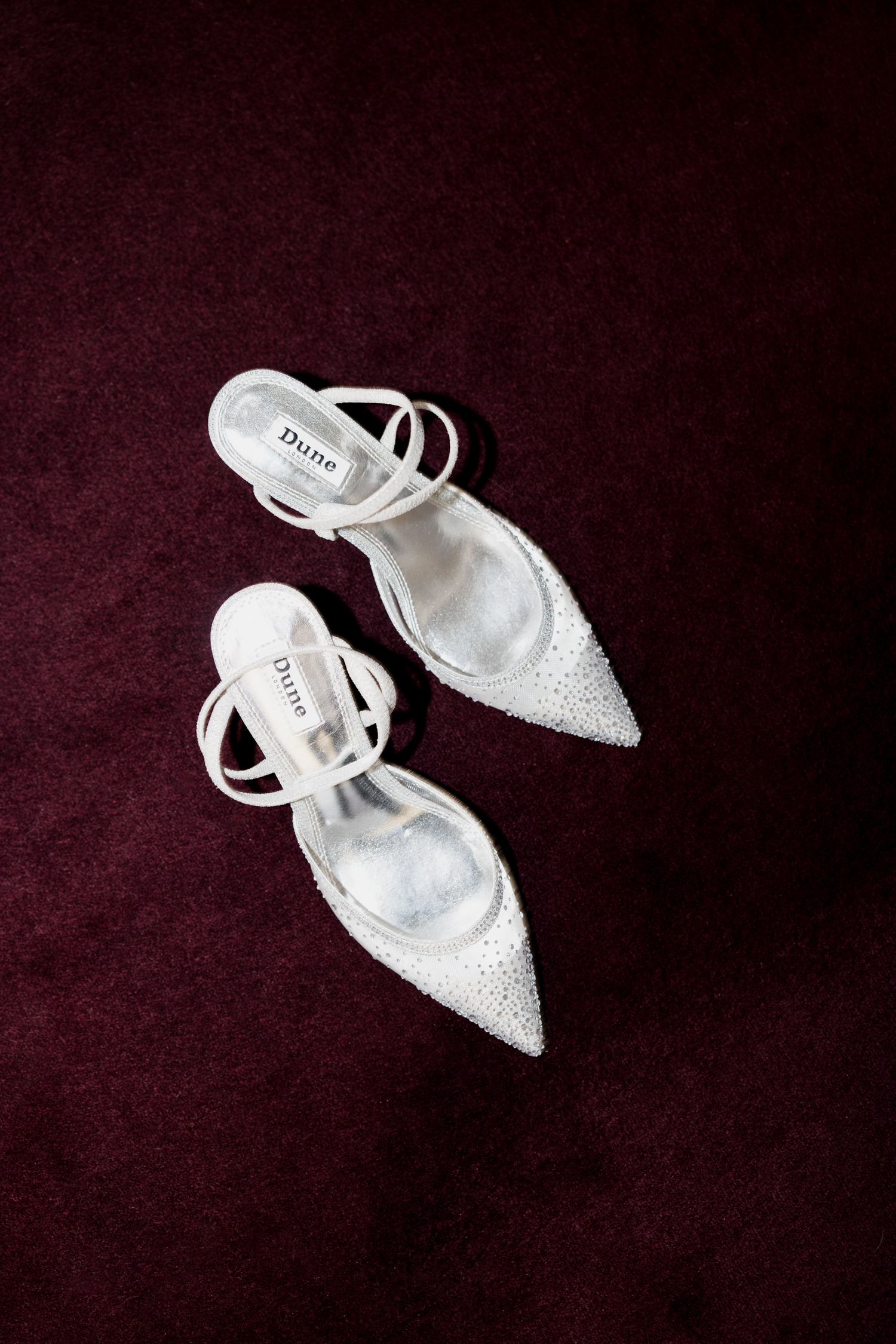 Elegant white glitter bridal shoes with pointed toes and ankle straps displayed on burgundy velvet background. Paris wedding photographer