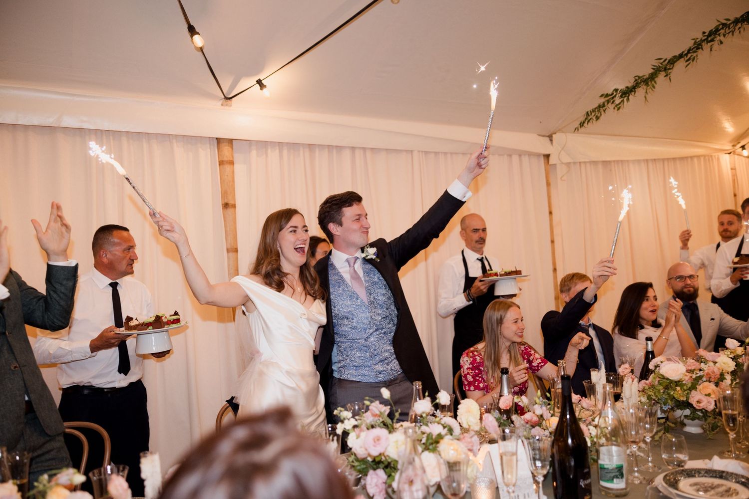 Wedding guests celebrate with raised arms during a reception dinner in a decorated white tent with twinkling lights.