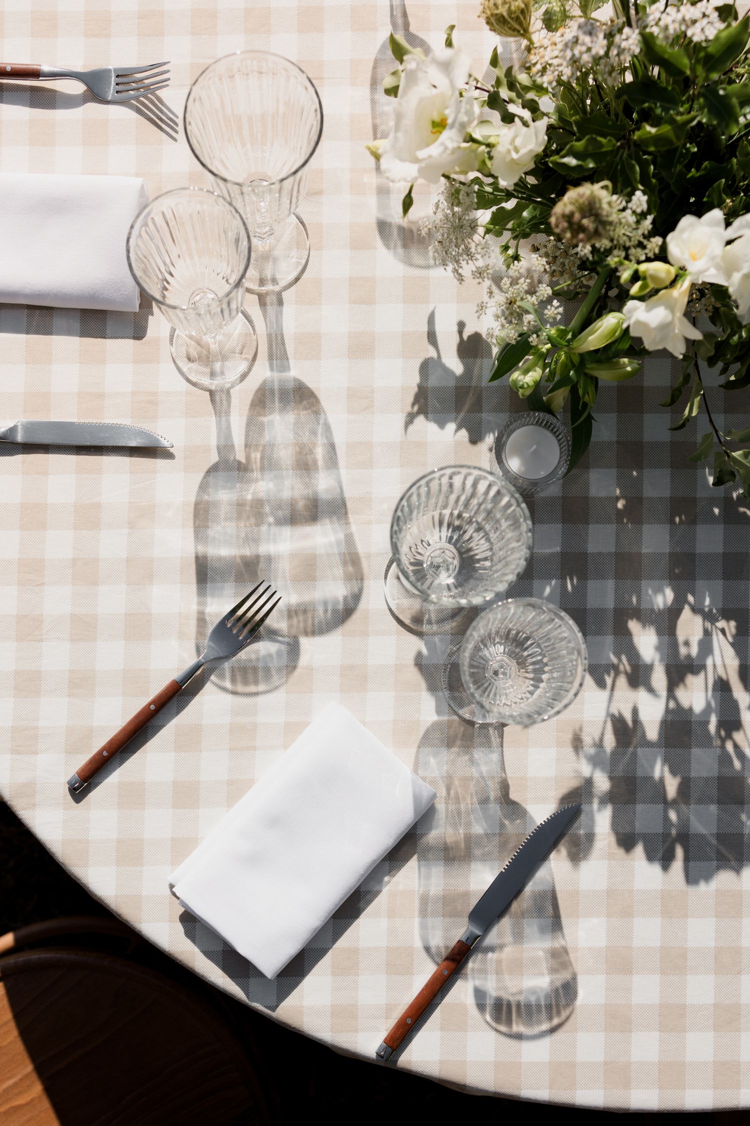 Elegant table setting with crystal glasses and white flowers on checkered tablecloth casting dramatic shadows.