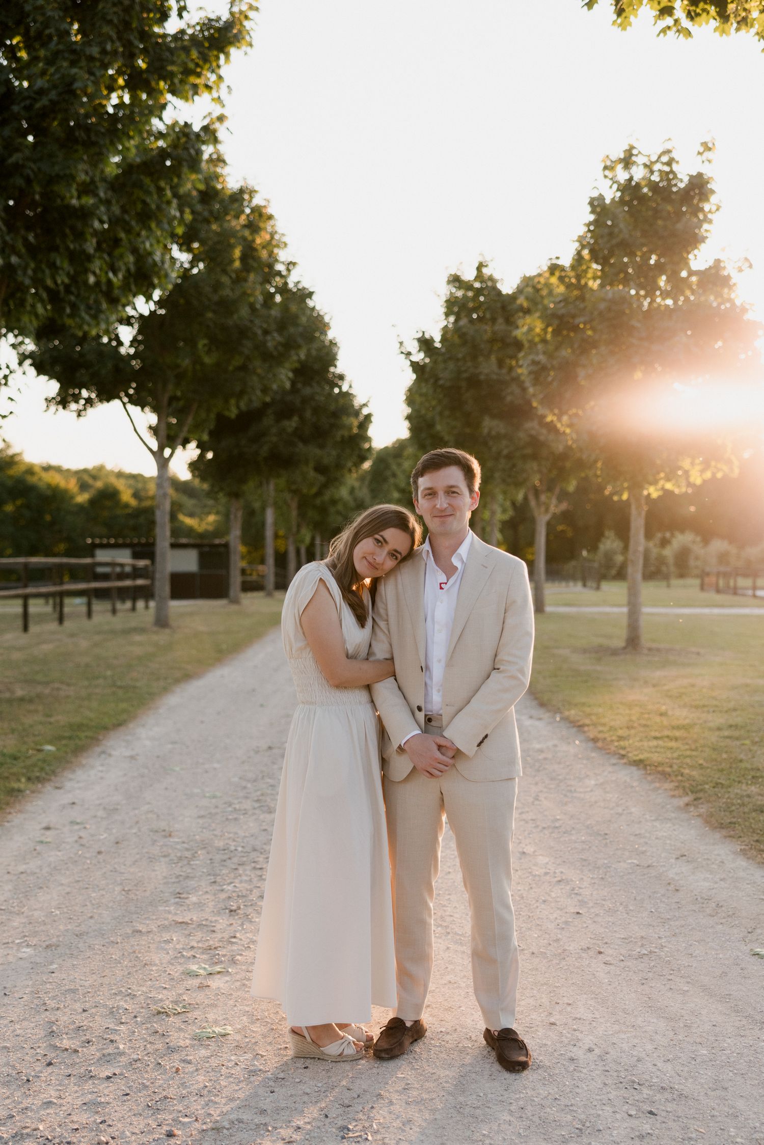 A couple in wedding attire walks along a sunlit dirt path surrounded by trees during golden hour.