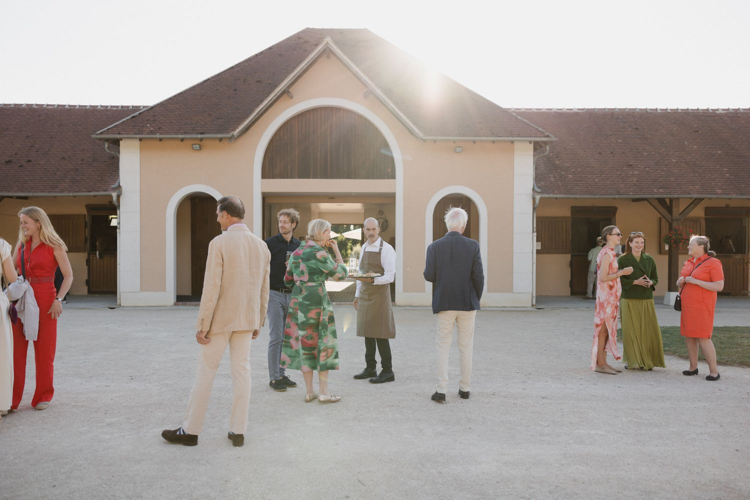 A group of people stand in a line outside a beige building with arched windows during a gathering event.