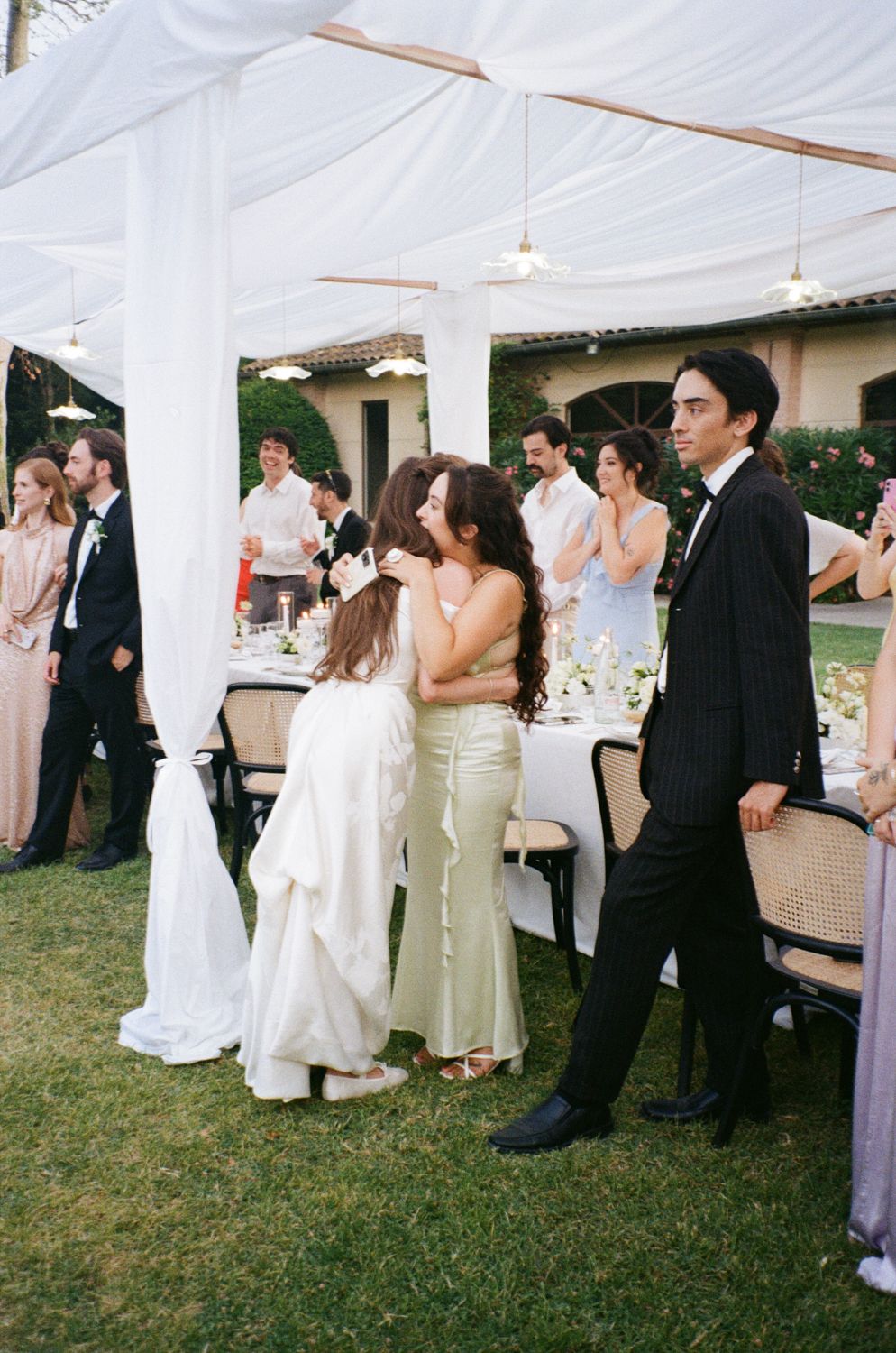 Wedding guests socialize under white tent canopies during an outdoor reception on a grassy lawn.