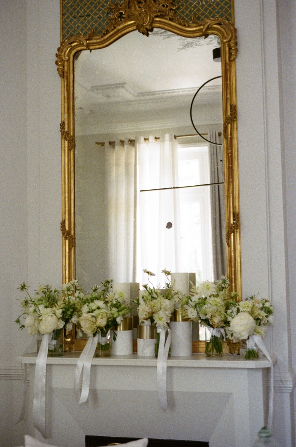 Ornate gold-framed mirrors line a white wall with delicate floral arrangements displayed on a ledge below.