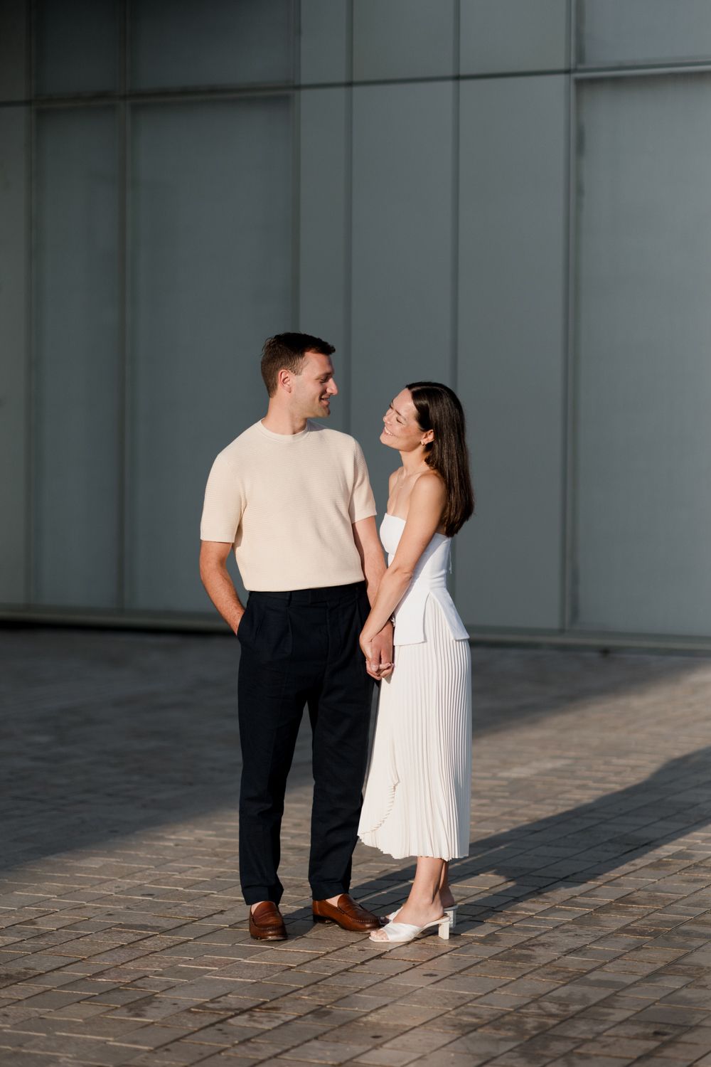 A couple in casual attire stands together on cobblestone pavement in front of an industrial grey wall. Photographe mariage Tours, France