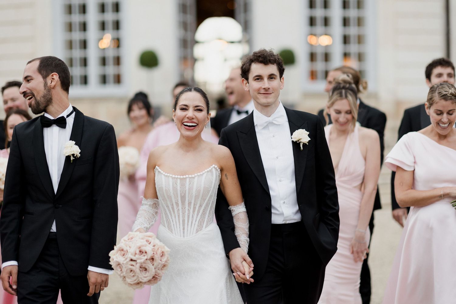 A smiling bride in a white corset gown holds pink flowers beside her groom in a tuxedo, with wedding party in pink and black.