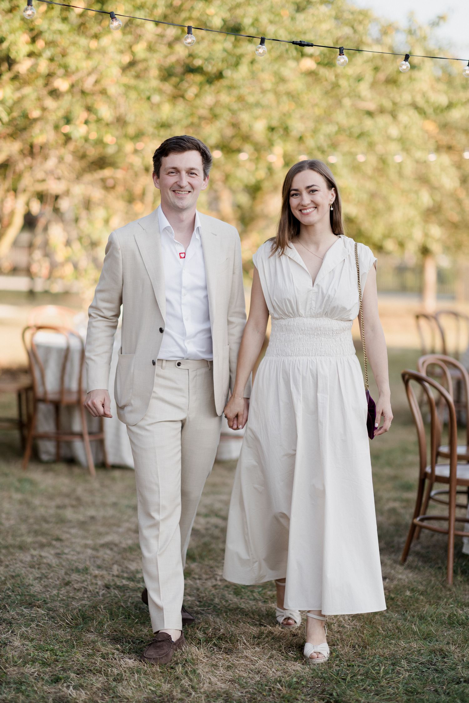 A couple in matching light-colored outfits walks hand in hand on grass during a rustic outdoor wedding celebration. Loire valley wedding pho