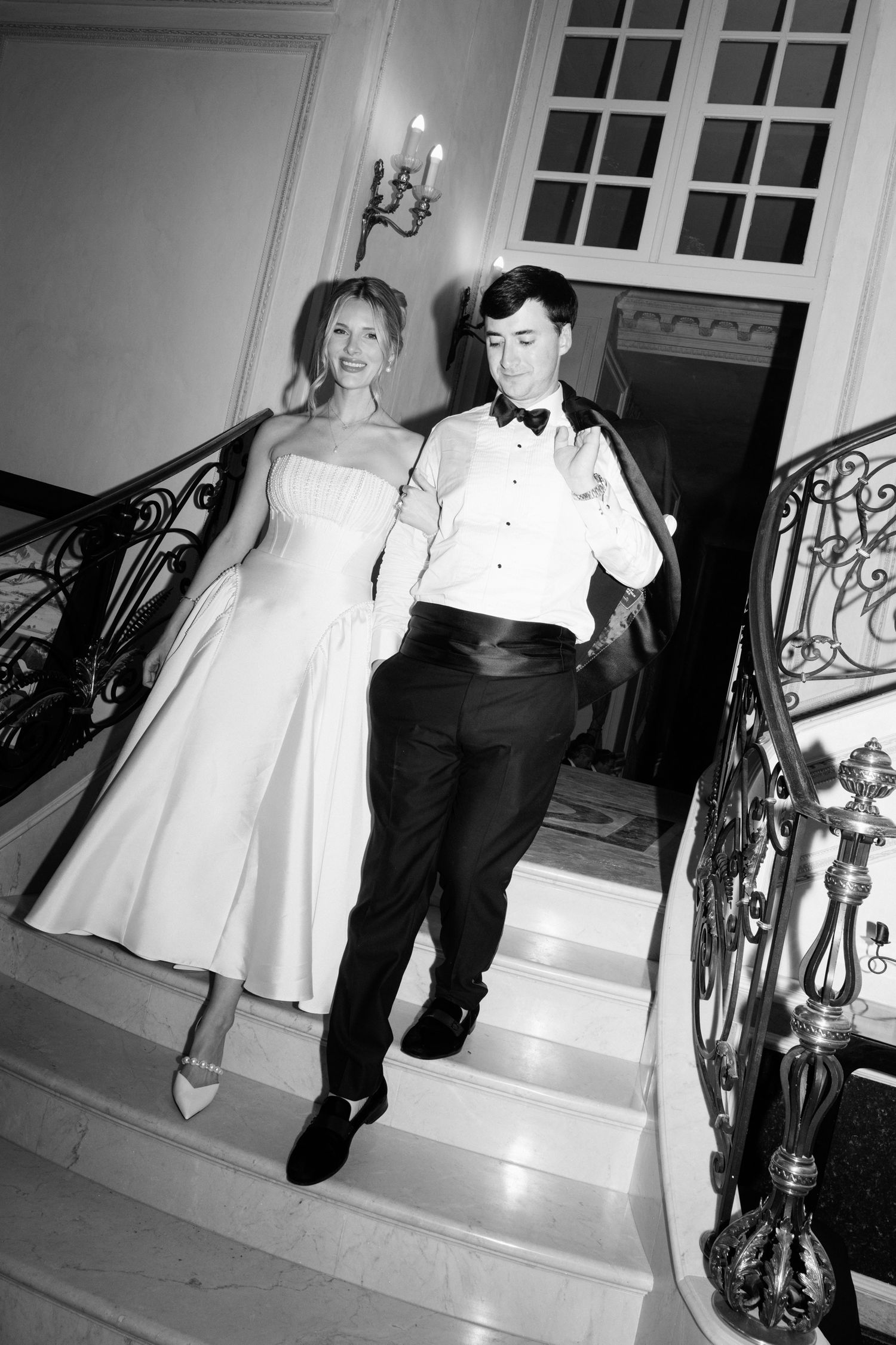 A couple descends an elegant staircase with ornate iron railings at what appears to be a formal event. Editorial wedding photographer france