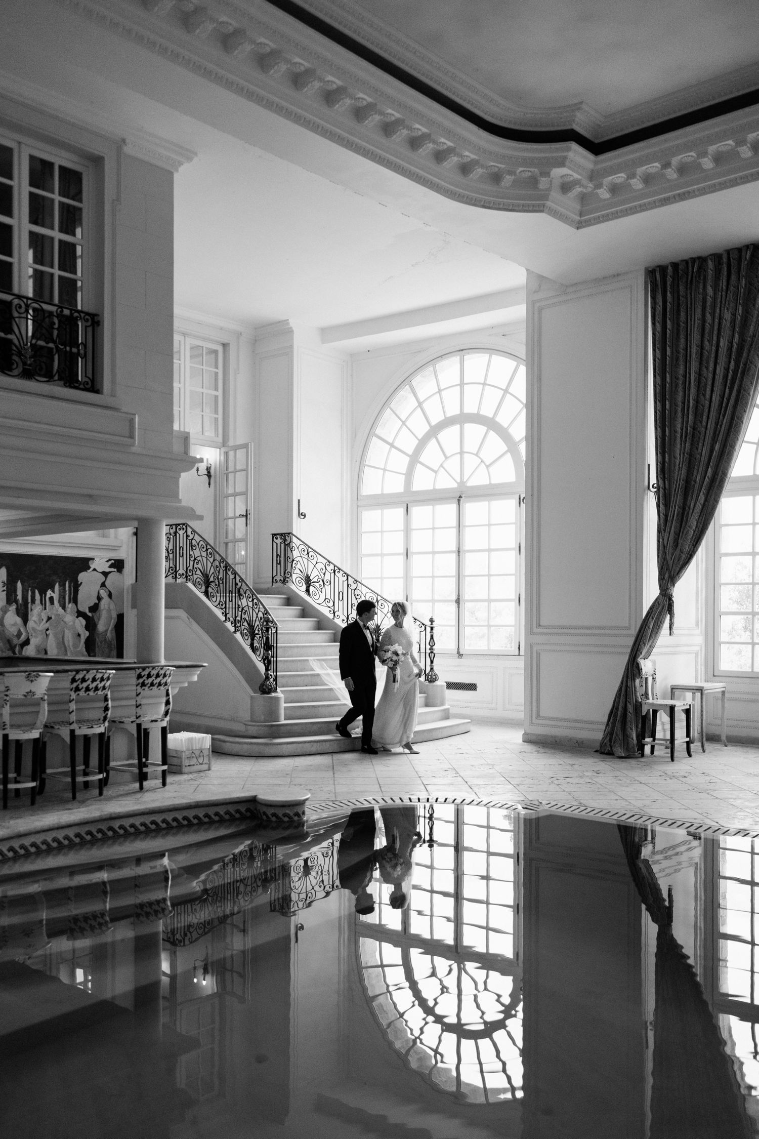 Elegant indoor swimming pool with ornate white moldings and arched windows reflecting on still water in black and white photography.