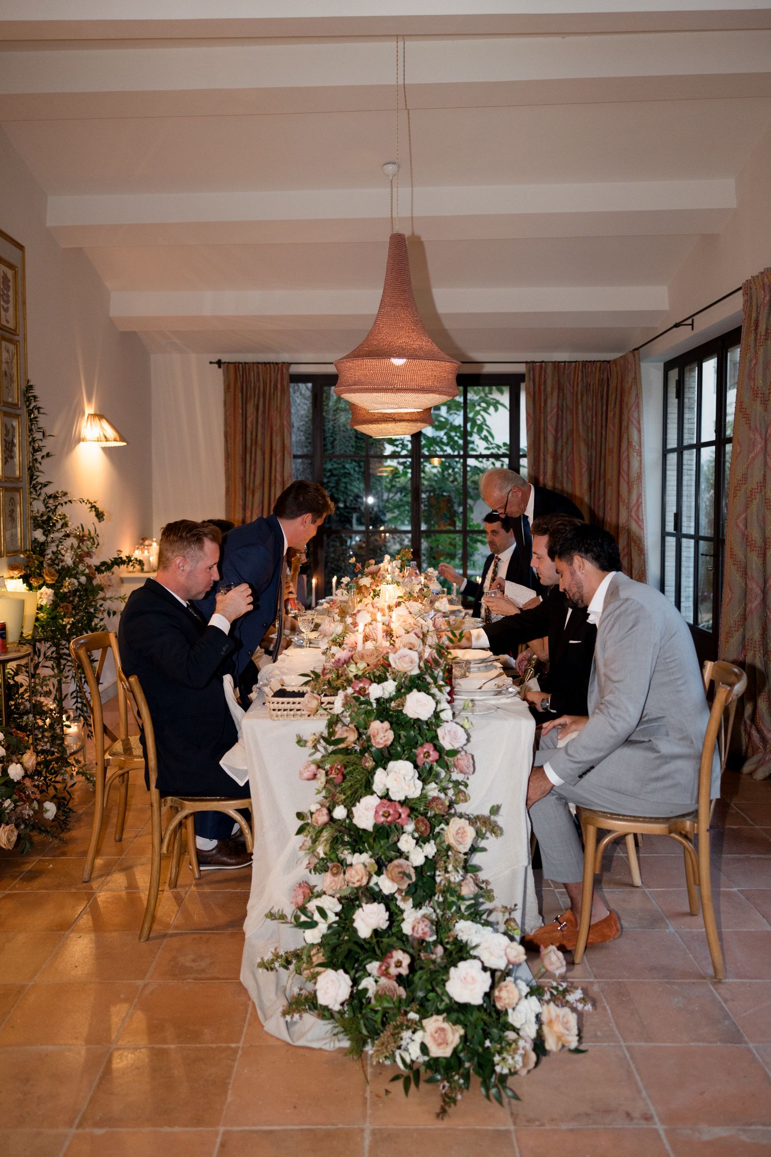 Elegant wedding reception dinner setup with floral centerpieces and guests seated at long white tables in a bright room.