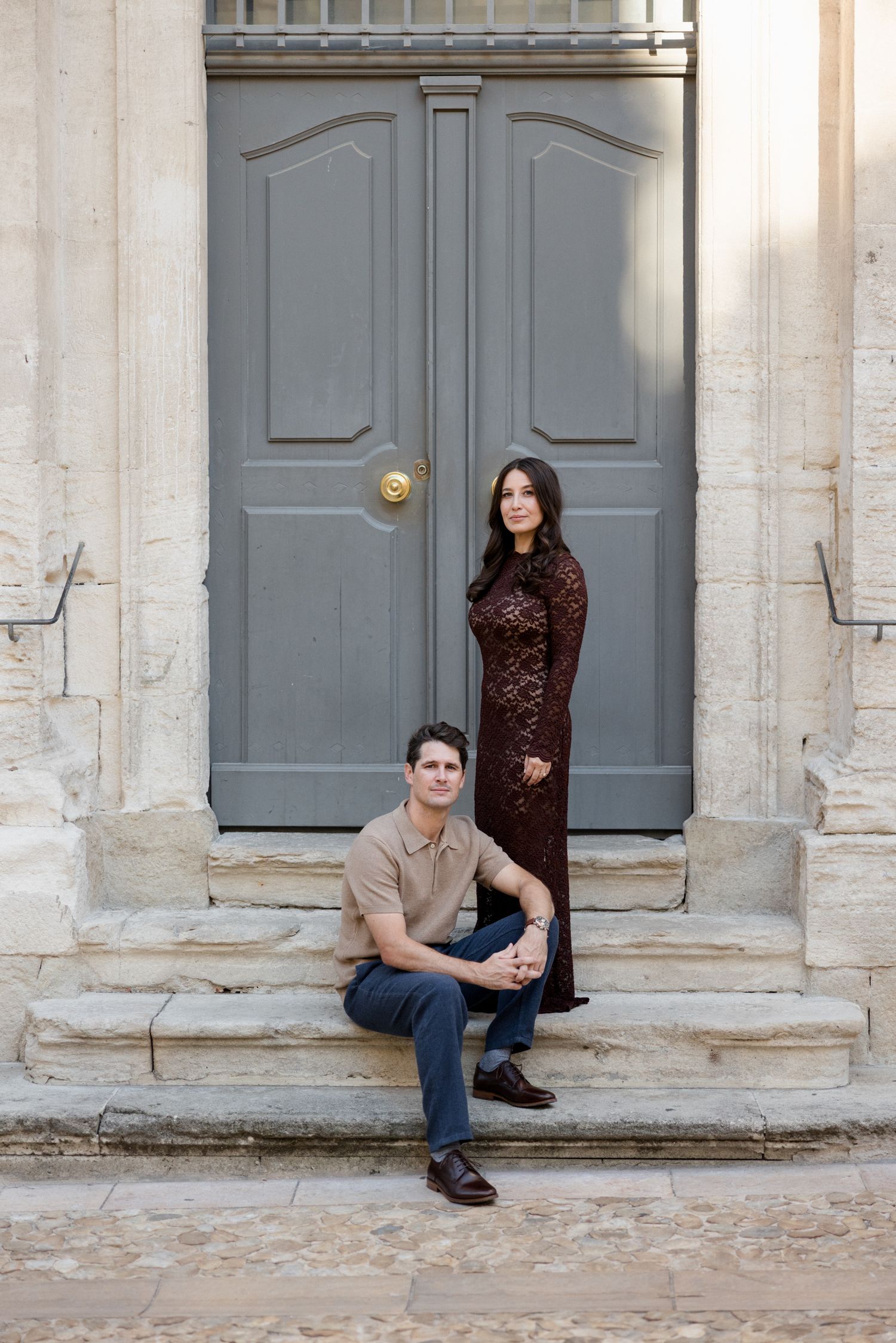 A couple poses casually in front of a gray door and white stone columns in an architectural setting. Engagement session France
