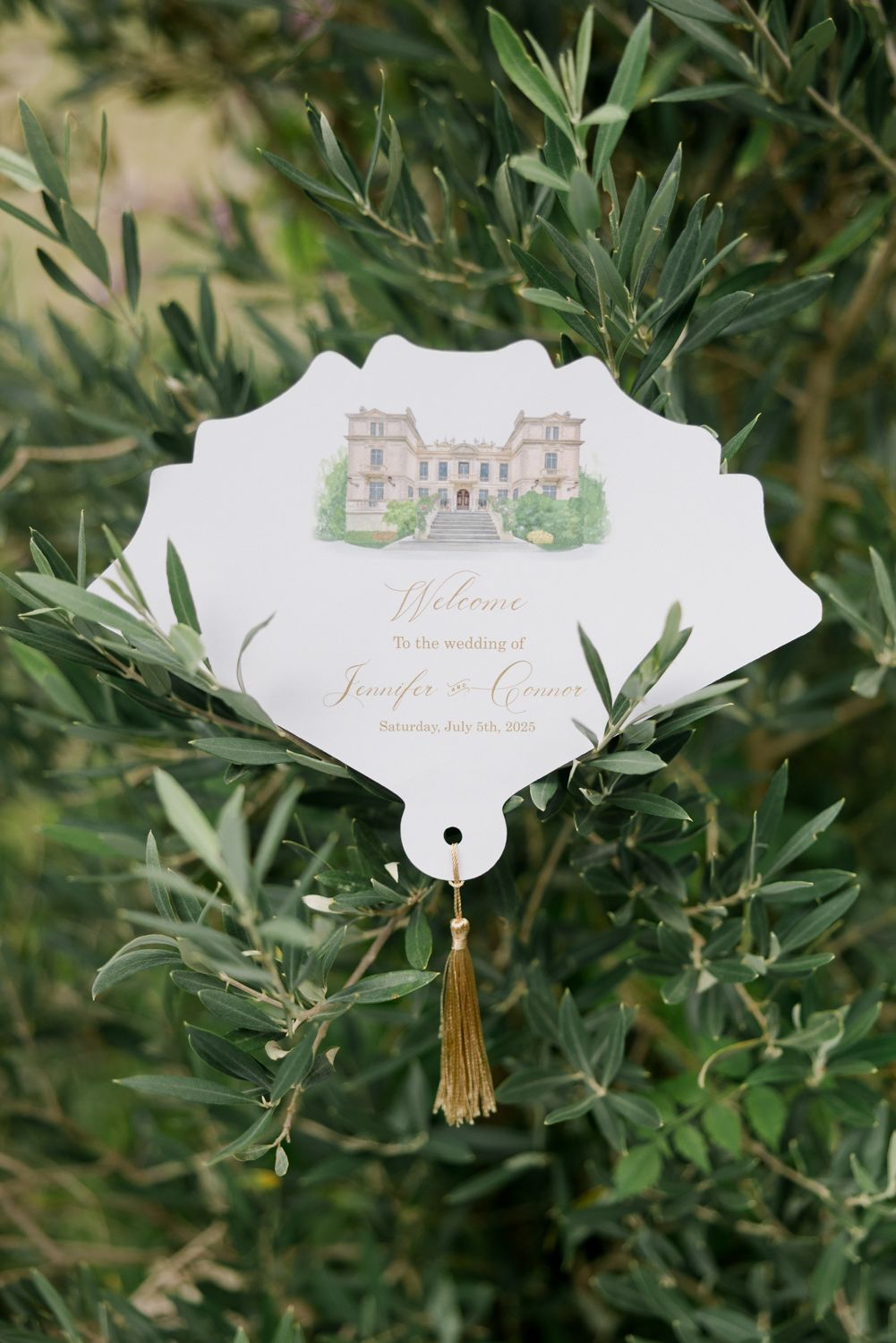 White paper wedding program fans adorned with calligraphy text displayed against green olive tree foliage.