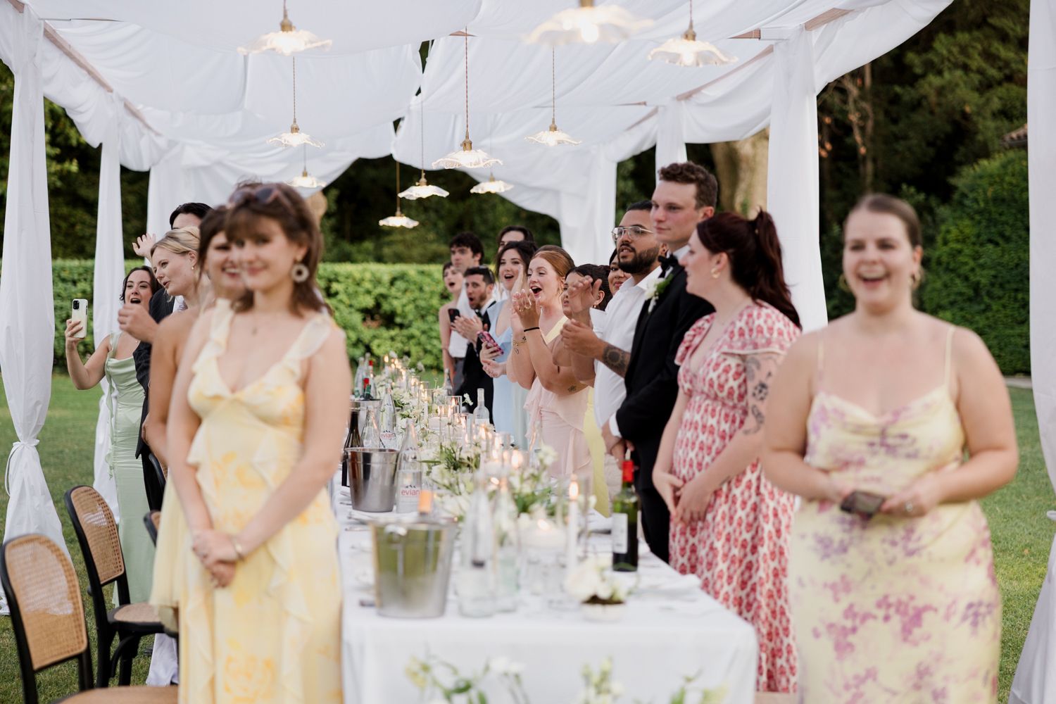 Guests gather under a white tent canopy at an elegant outdoor garden reception with table settings and string lights.
