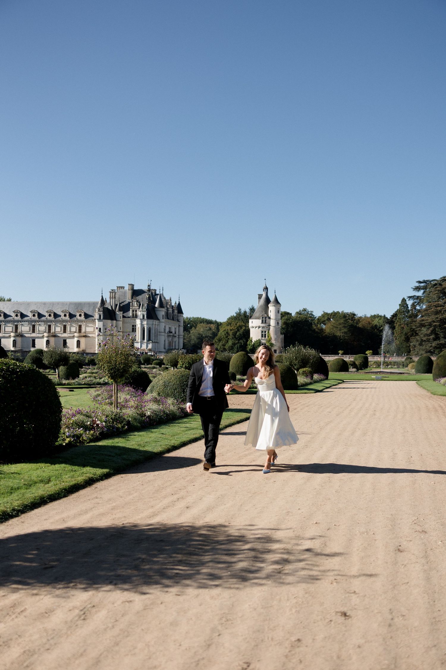 Bride and groom walking hand in hand along a gravel path with a grand French castle behind.