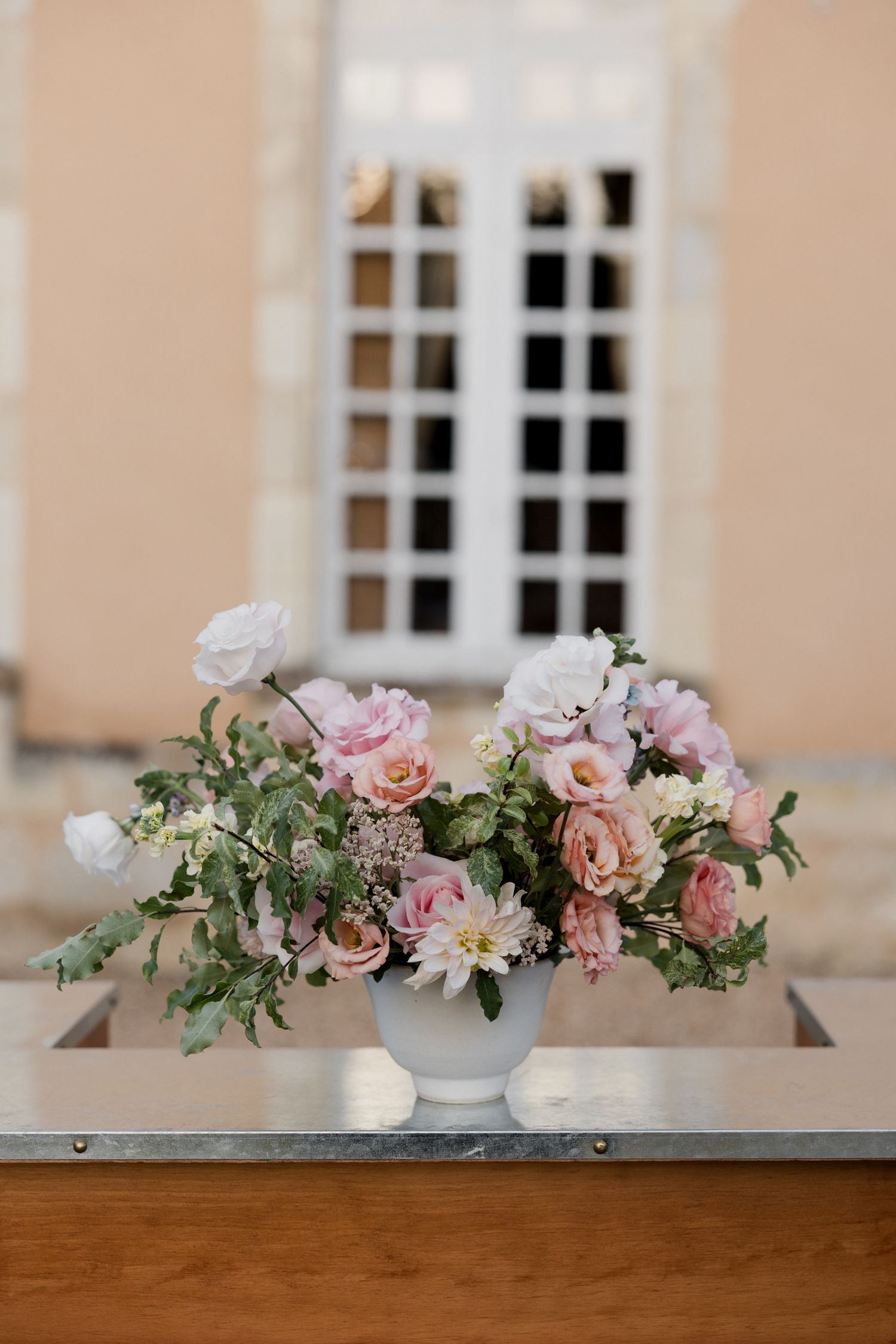 Row of pink and peach floral centerpieces arranged on wooden surface against cream-colored wall with windows.