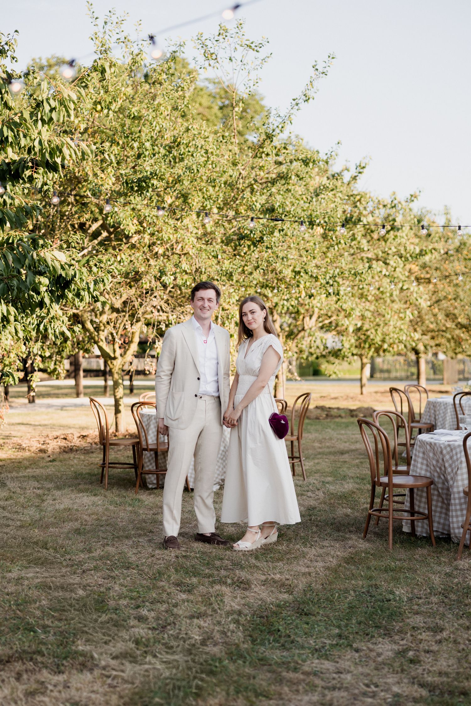 A couple stands together at an outdoor garden wedding reception with wooden chairs and blooming trees in the background. Loire valley weddin