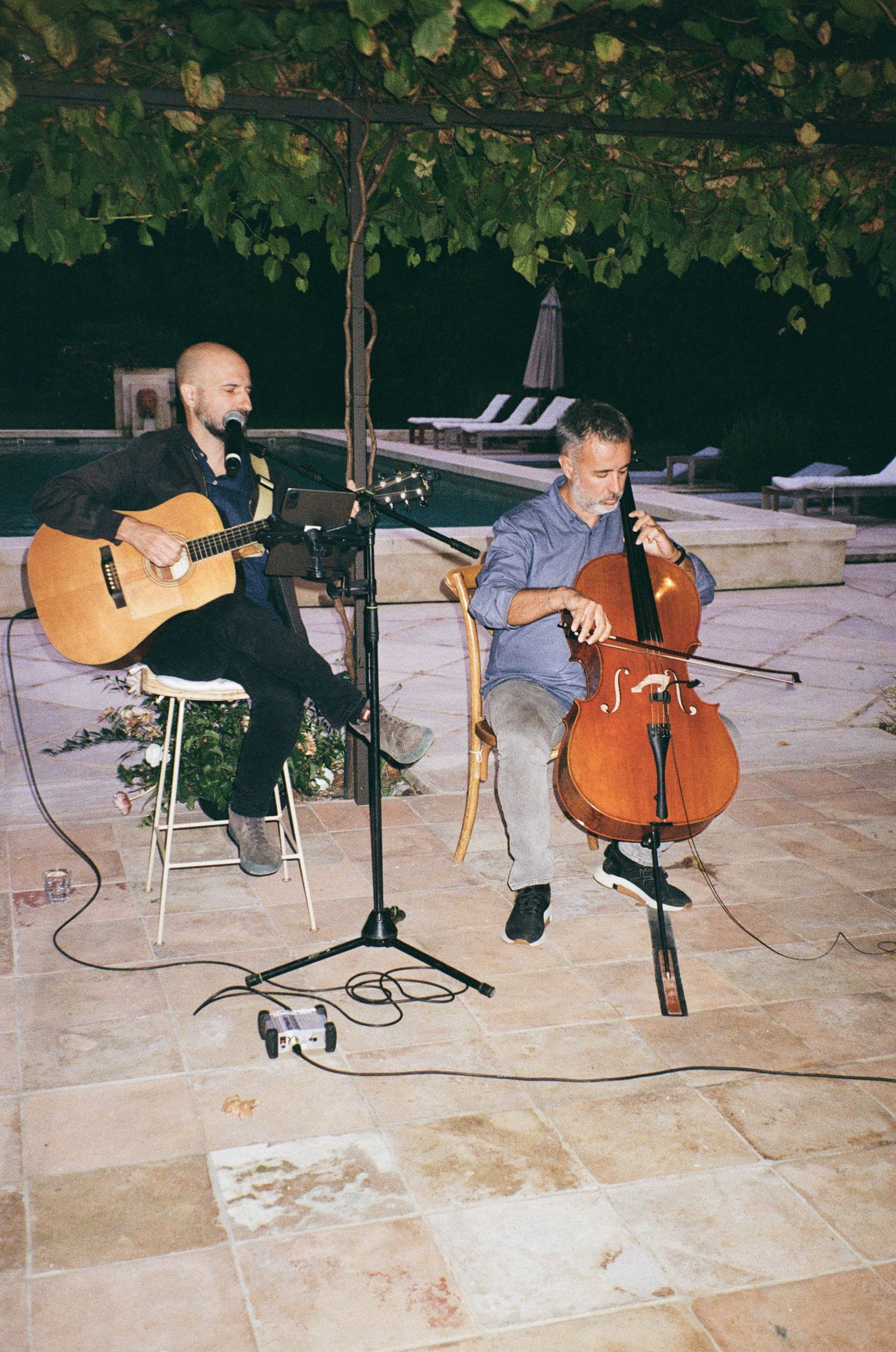 Musicians perform with acoustic instruments including guitar and cello on an outdoor stone patio under leafy tree cover.