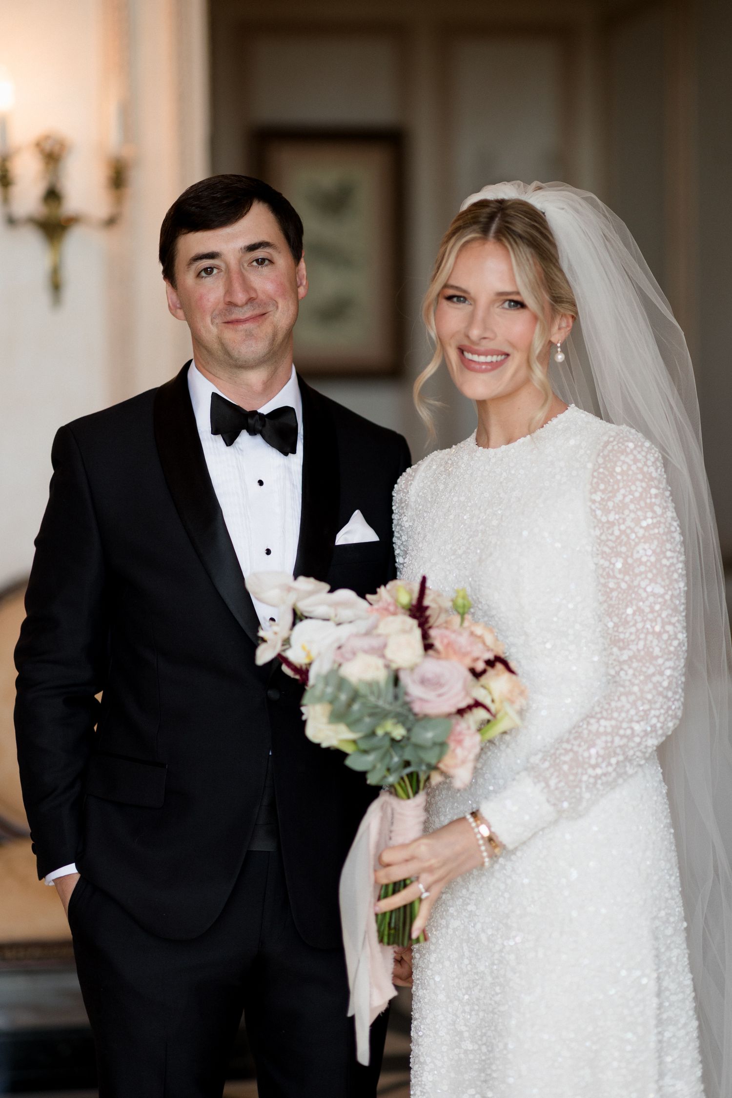 A bride in a long-sleeved lace wedding dress and groom in black tuxedo pose for portraits in an elegant venue. editorial wedding photographe