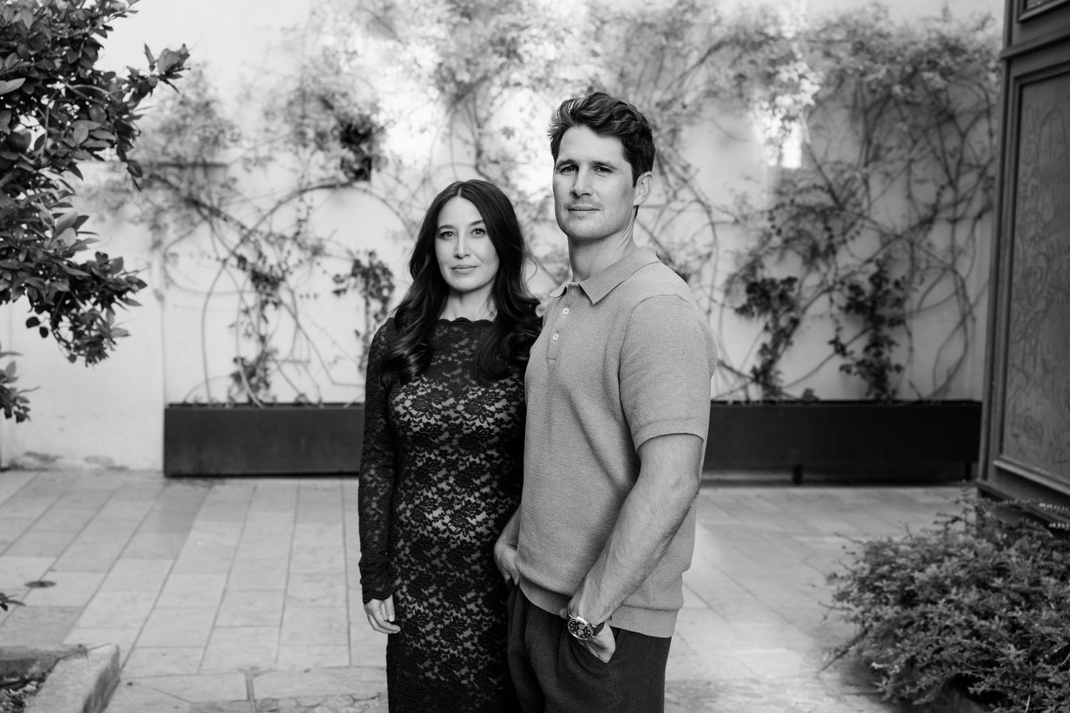 Black and white portrait of a couple standing together against a textured wall with climbing vines.