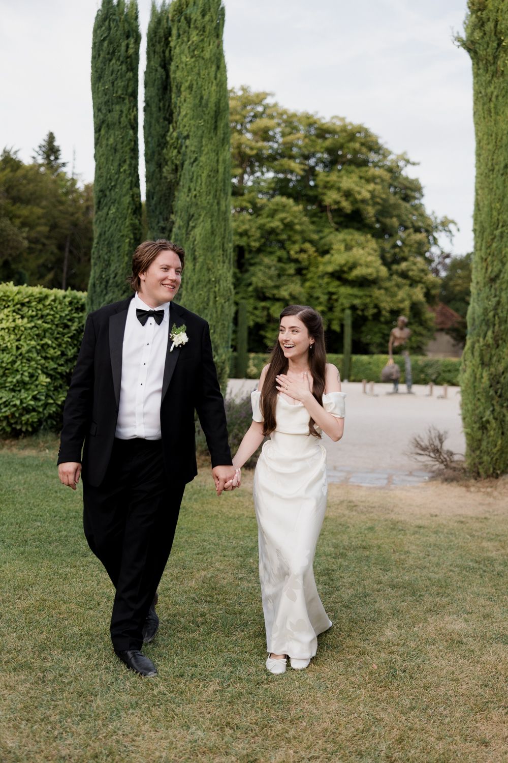 A couple in formal wedding attire walks and laughs together in a garden setting with tall cypress trees.