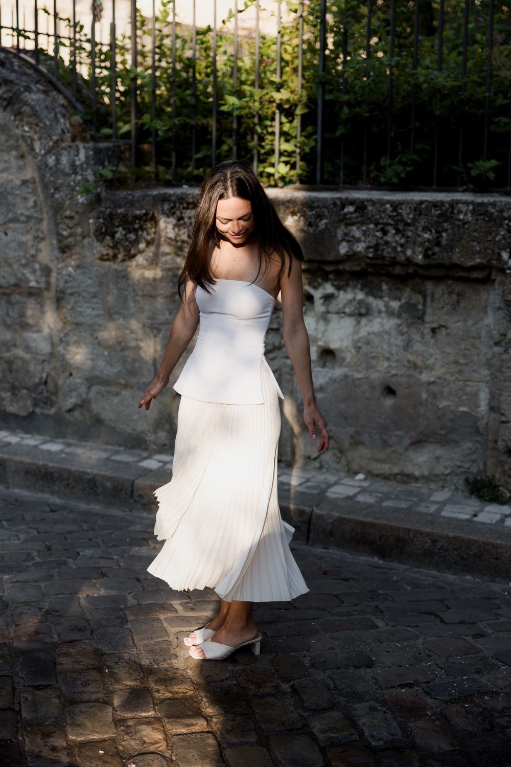 A person in a flowing white dress twirls on a cobblestone path against a weathered stone wall at dusk.