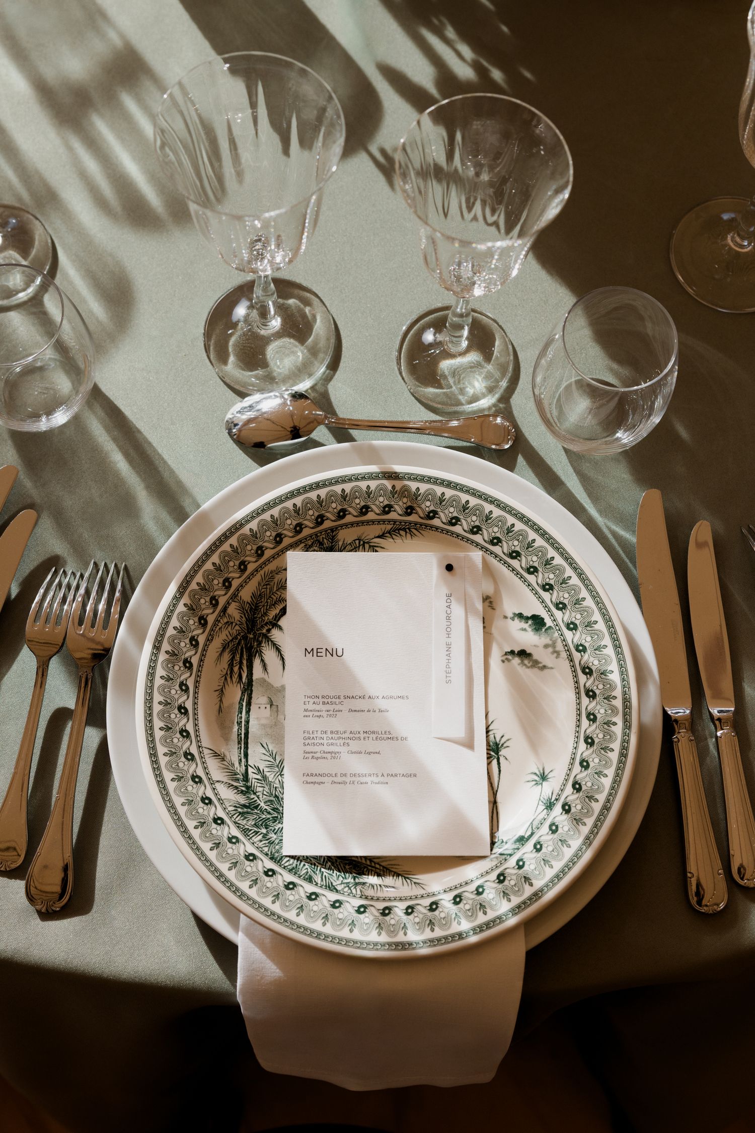 Elegant place setting with patterned china plate, menu card, crystal glasses and vintage cutlery on white tablecloth.