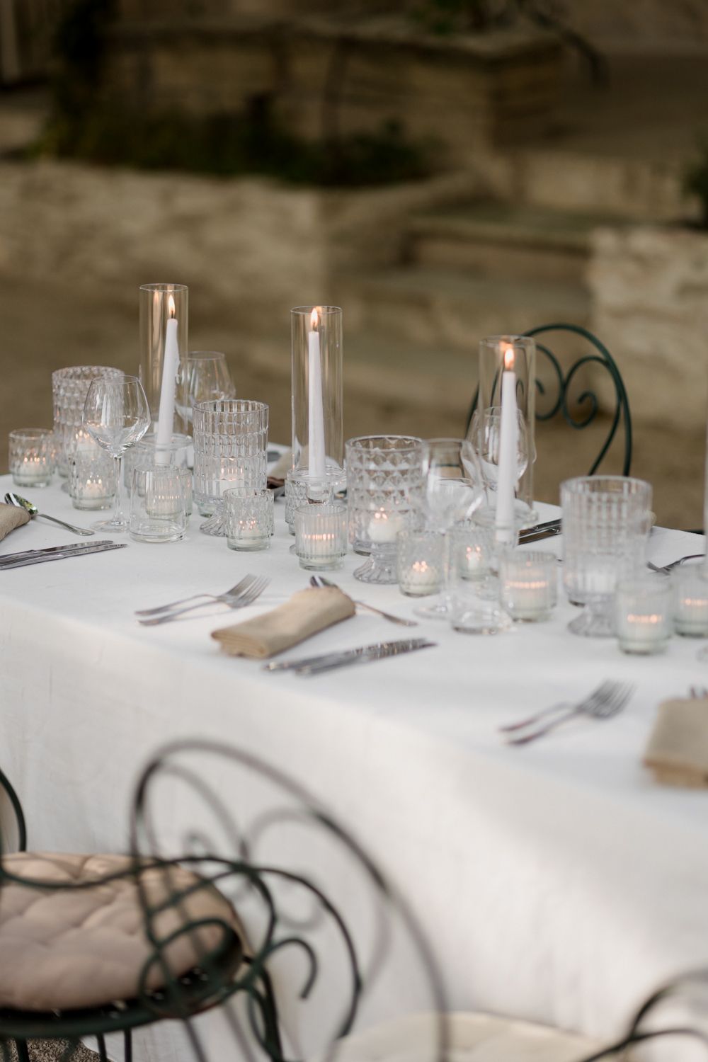 Romantic dinner table with crystal glassware, tea lights and wrought iron chairs set against rustic background. Auberge de la Treille