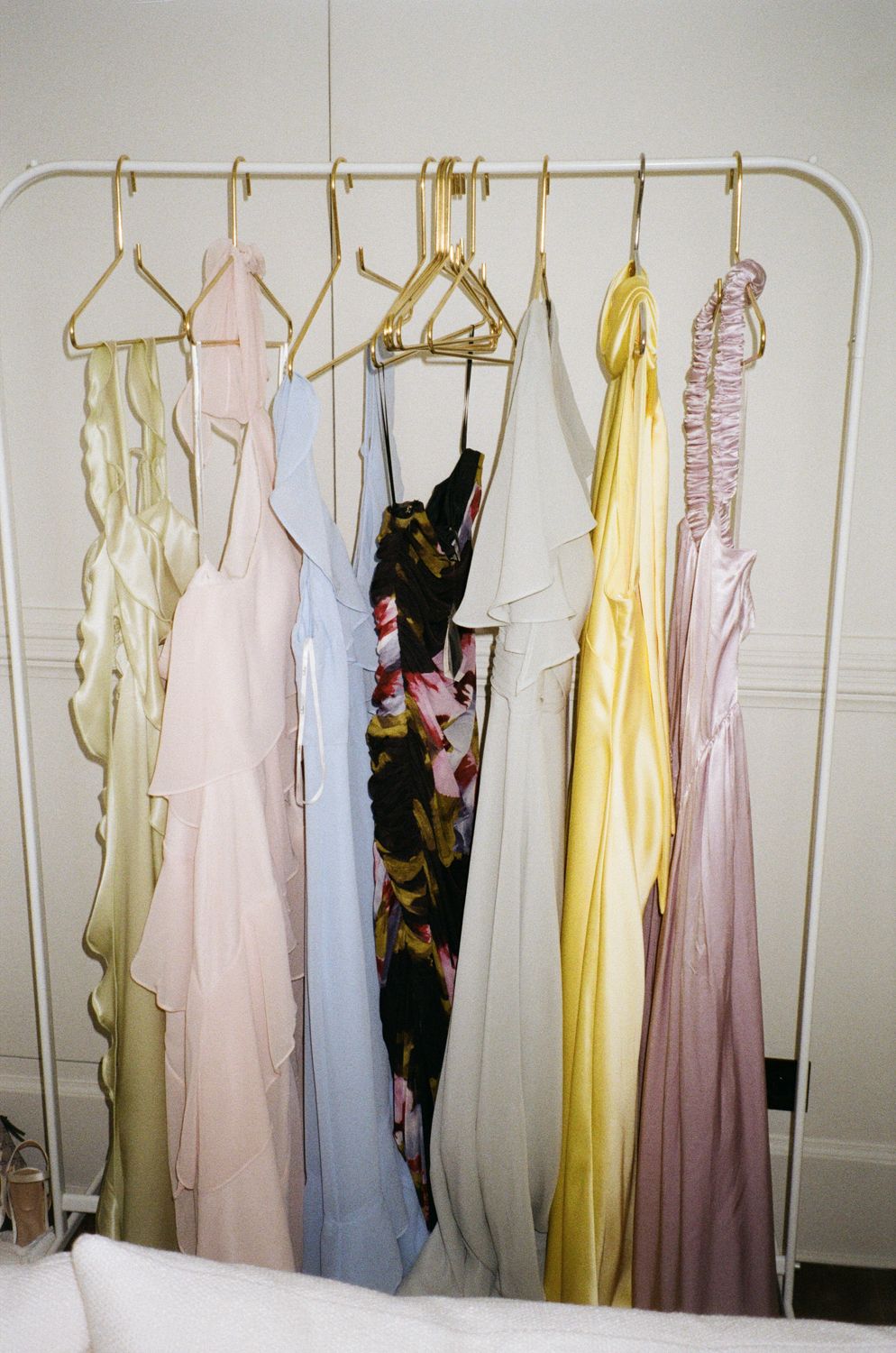 Vintage dresses in soft pastel colors hanging on gold hangers against a white wall, showing delicate formal evening wear.