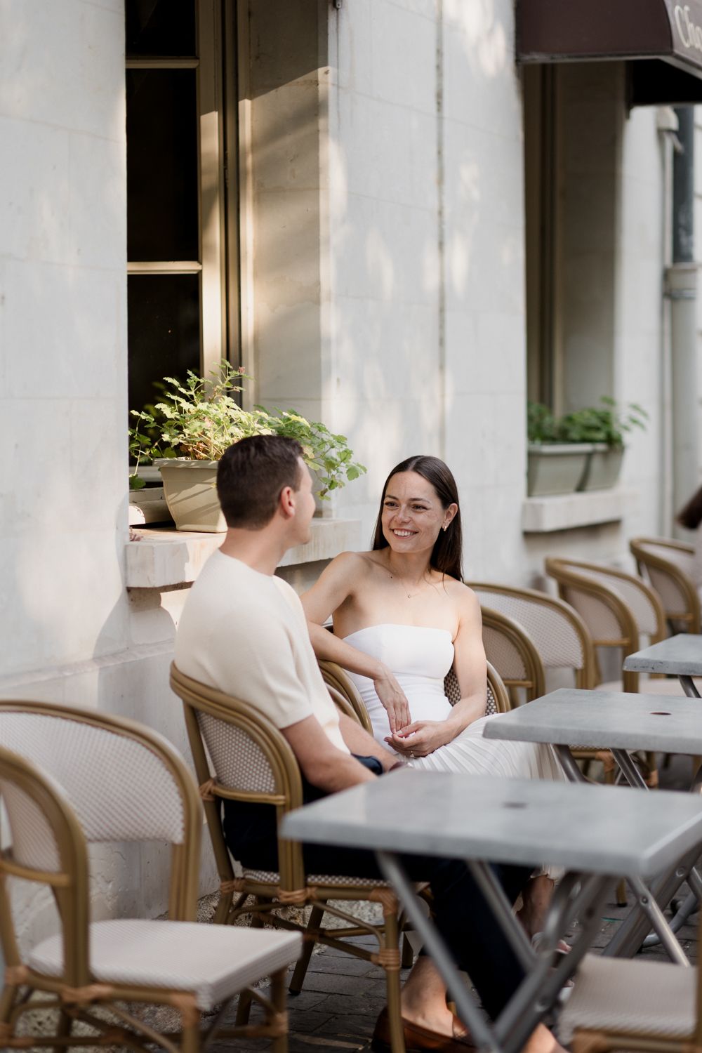 Couple having an intimate conversation at an outdoor cafe with white chairs and tables in natural lighting.