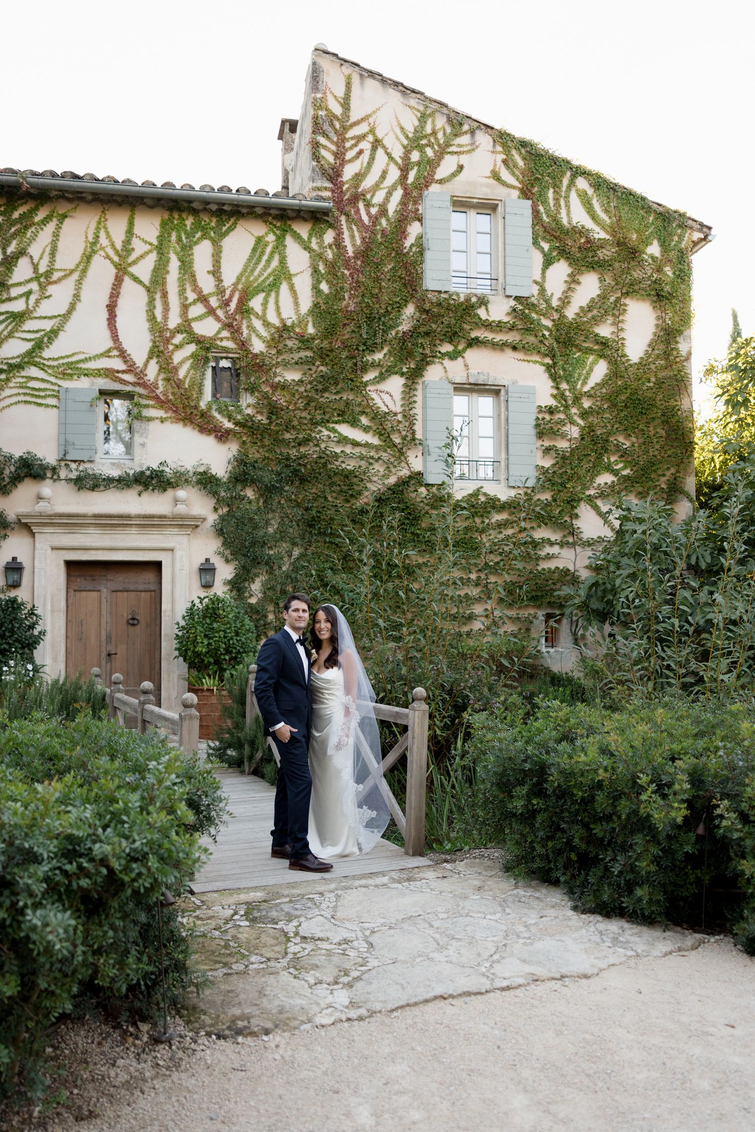 Romantic wedding couple poses in front of a rustic stone building with climbing vines and greenery. wedding photographer france
