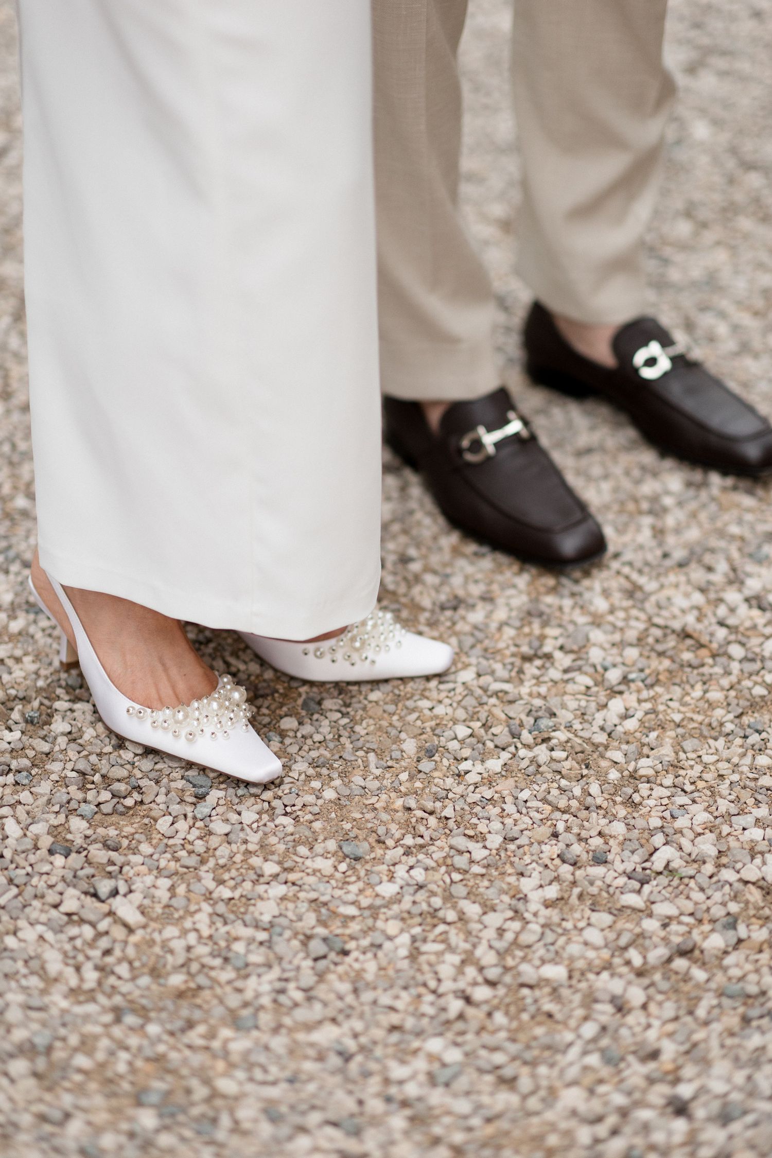 Close-up of elegant wedding shoes showing a white bridal flat and black groom's loafer on gravel ground.
