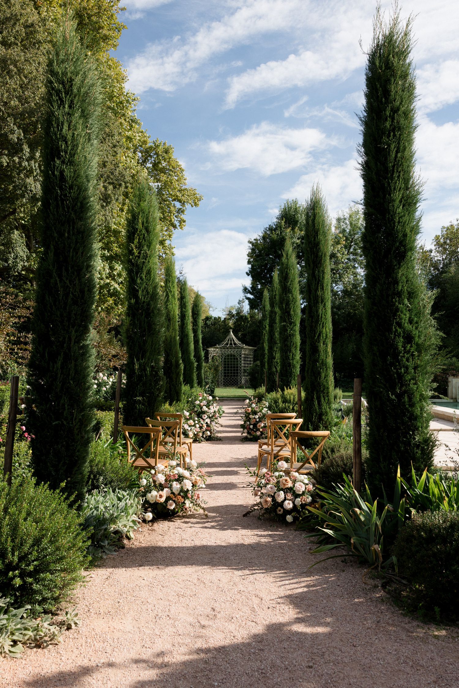 Garden pathway lined with tall cypress trees and flower beds leading to a gazebo under blue skies.