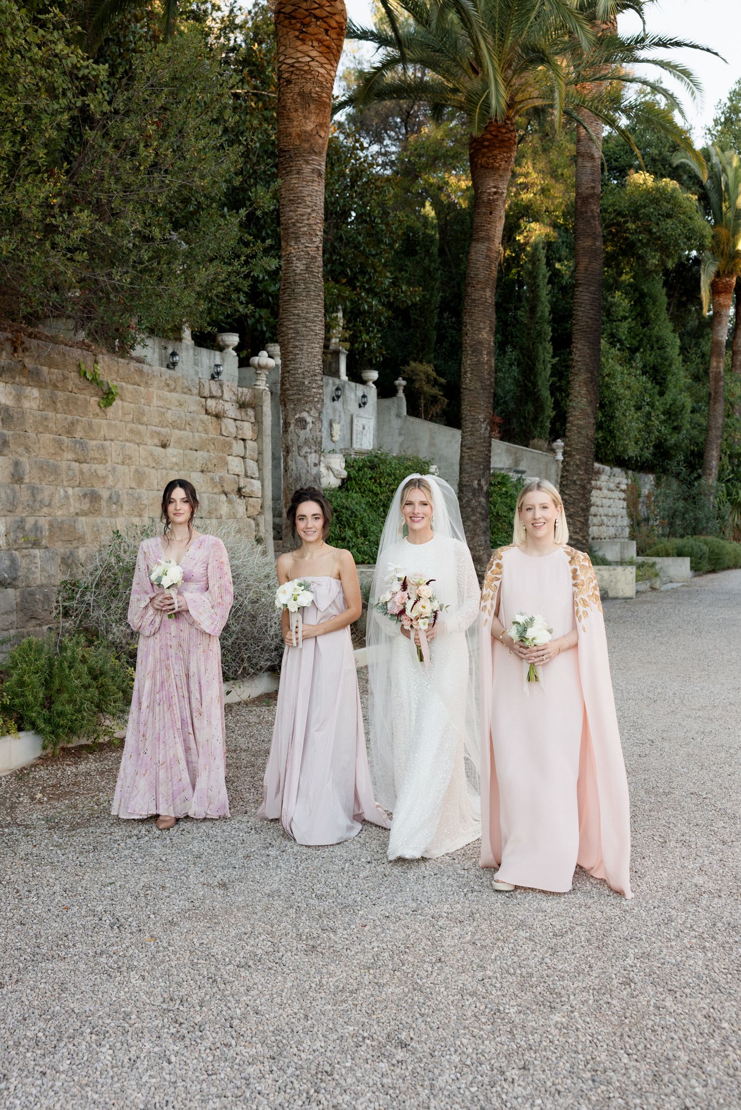Bridesmaids in soft pink dresses pose together on a palm tree-lined gravel path at an elegant venue. Photographe mariage france grasse azur