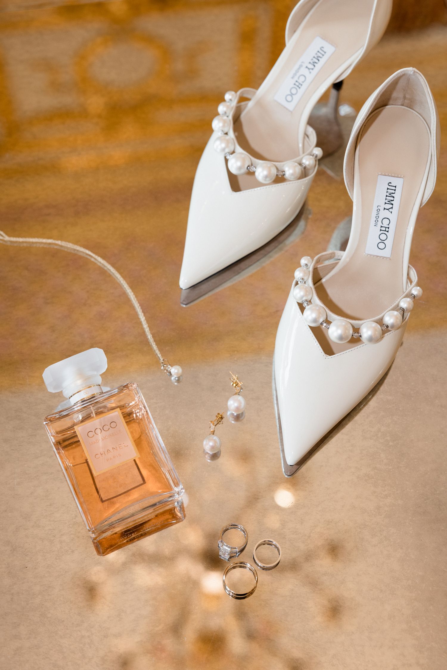 Elegant white Jimmy Choo heels with pearl details displayed next to perfume bottle and wedding rings on wooden surface. destination wedding