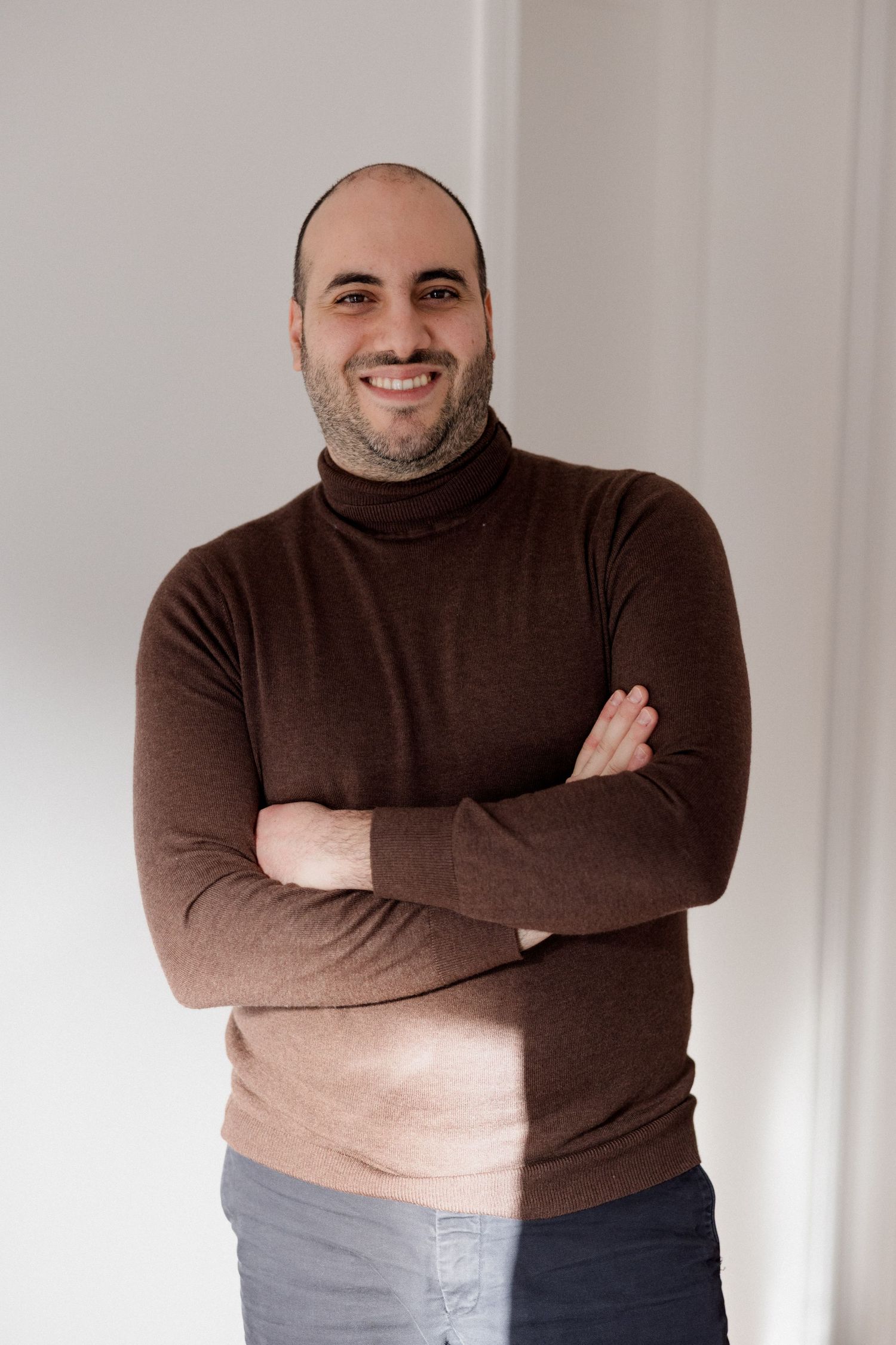 Smiling bald man with beard, arms crossed, wearing a brown turtleneck sweater, standing against a white wall.